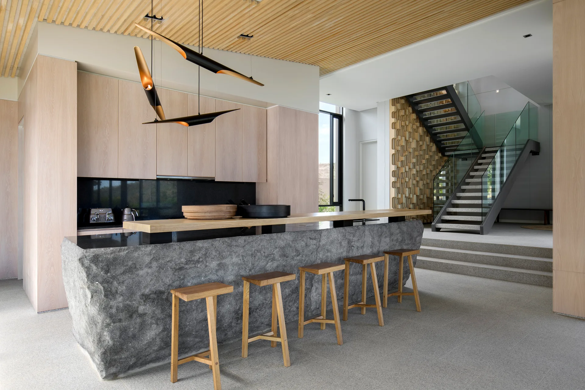 A modern kitchen featuring a large raw stone island with wooden bar stools under a slatted ceiling, with a staircase visible in the background.