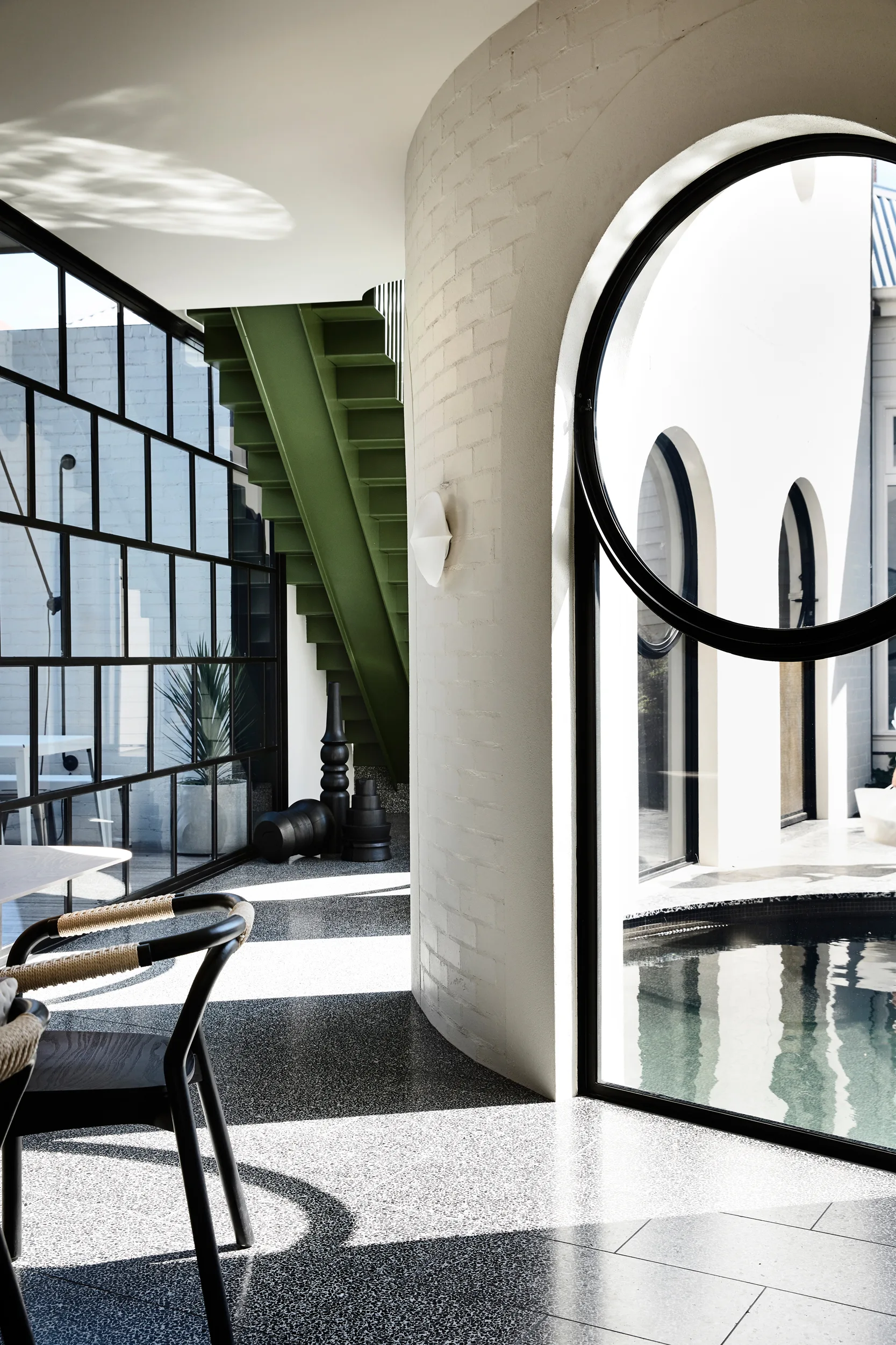 A modern interior featuring a curved white brick wall with an arched window overlooking a pool next to a green metal staircase.