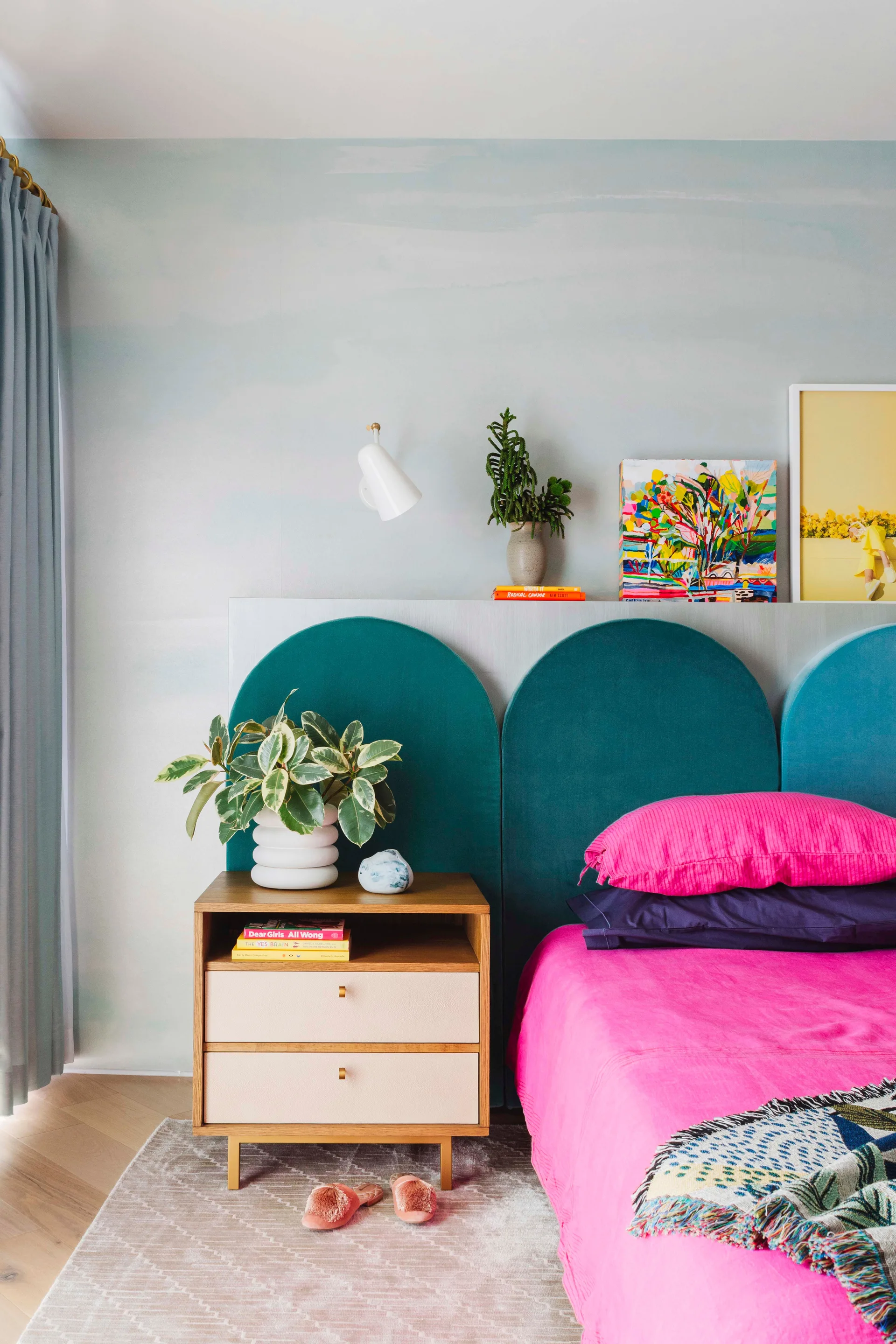 A modern bedroom features a segmented teal arched headboard, bright pink bedding, a wood nightstand, and a pale blue gradient accent wall.