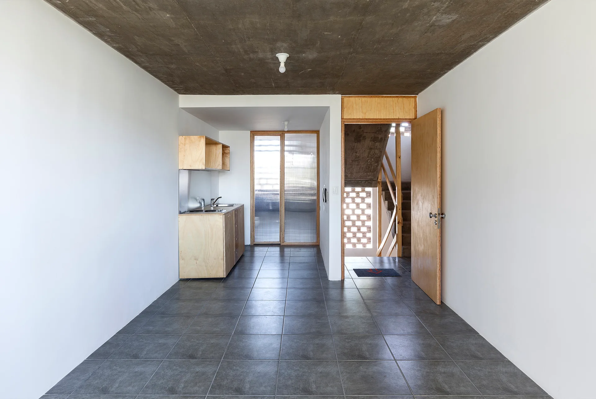 A minimalist kitchen interior with a concrete ceiling, grey tiled floor, plywood cabinetry, and a wooden door leading to a staircase.