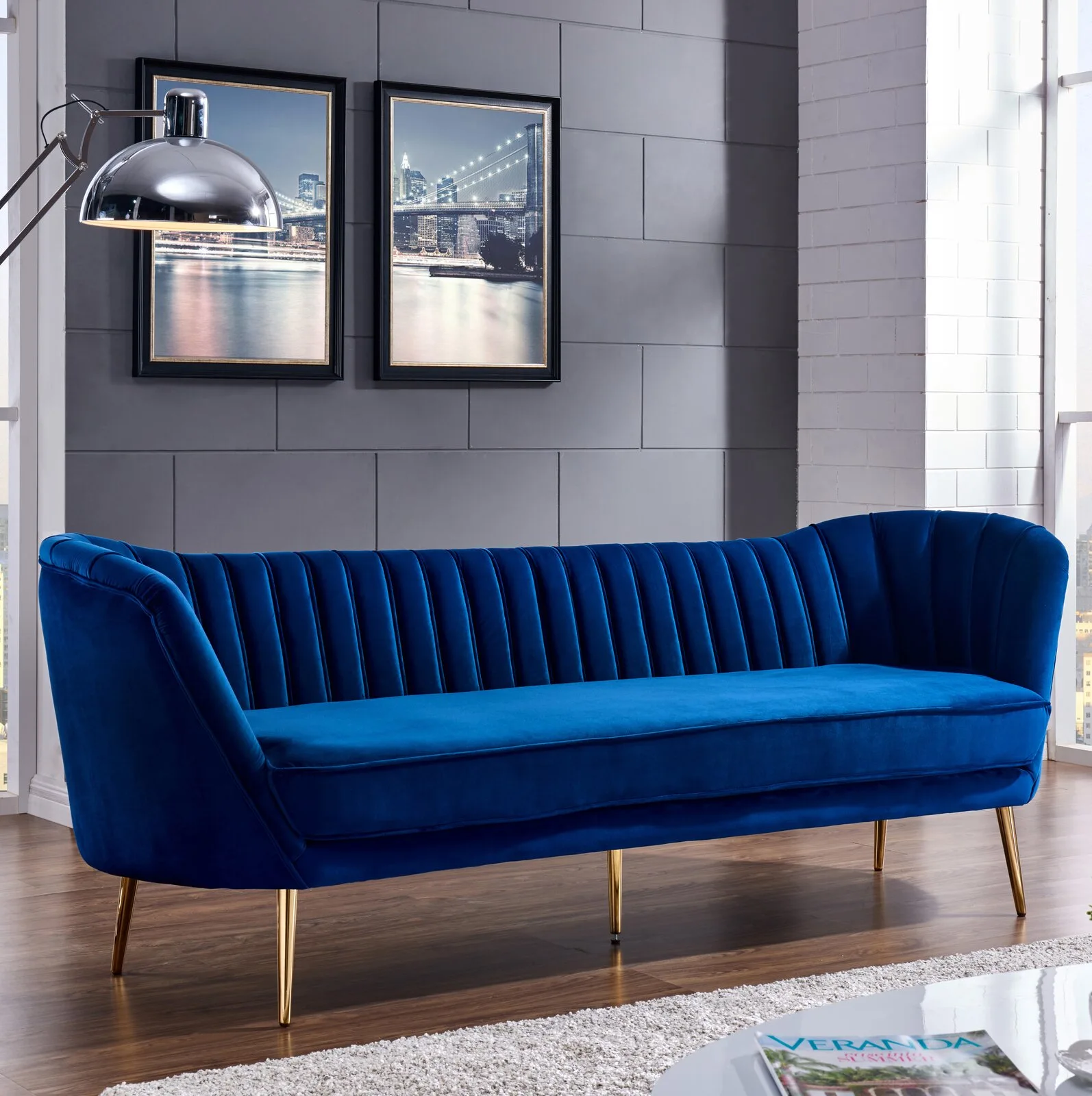 A vibrant blue velvet channel-tufted sofa sits on hardwood floors in front of a dark grey paneled wall featuring two framed cityscapes.