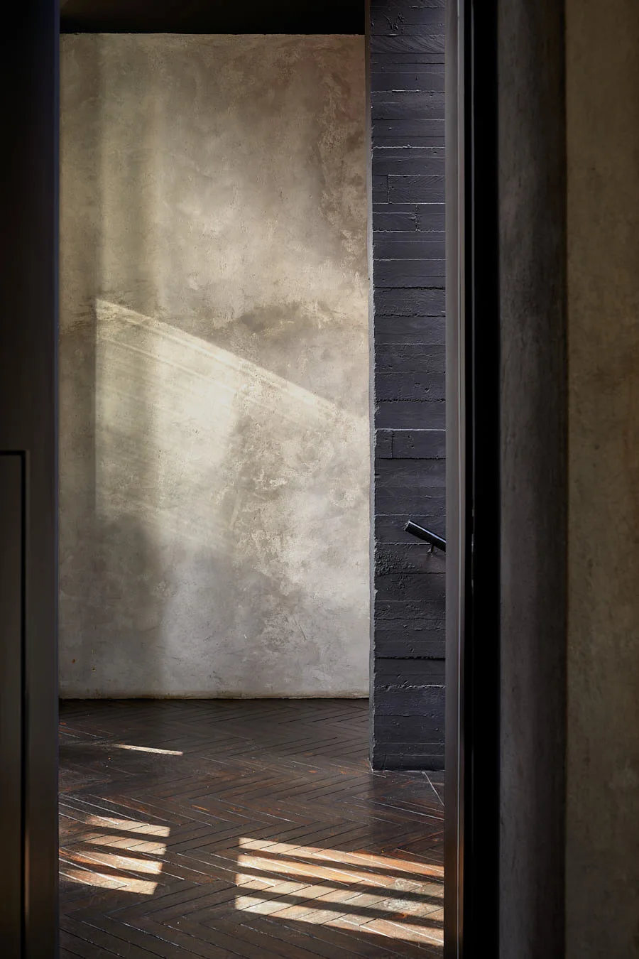 A doorway frames a view into a gray concrete hallway featuring a dark wood herringbone pattern floor and a textured black wall.