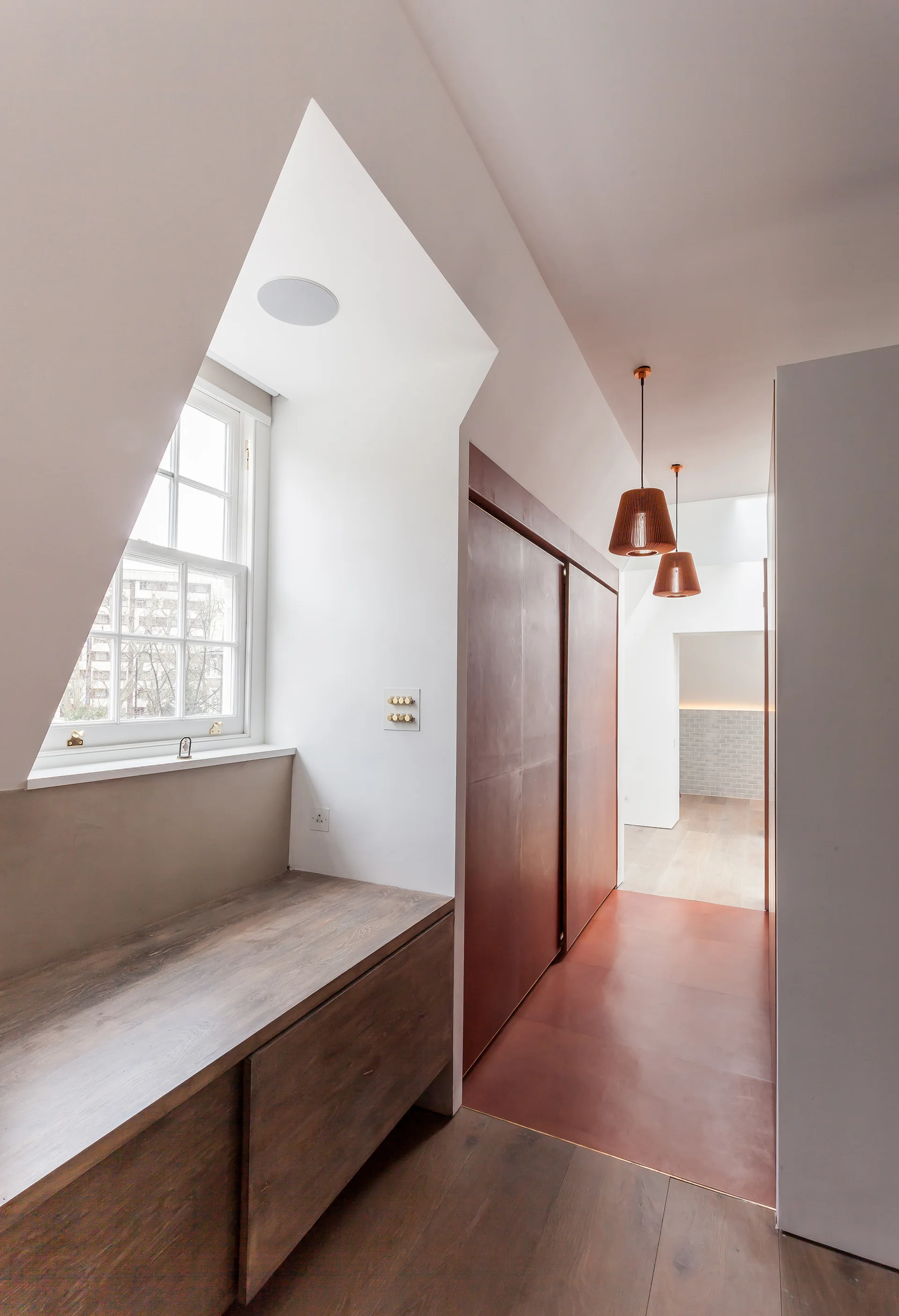 A white attic hallway features a built-in wooden bench below a window and tall copper-toned closet doors beneath two hanging pendant lights.