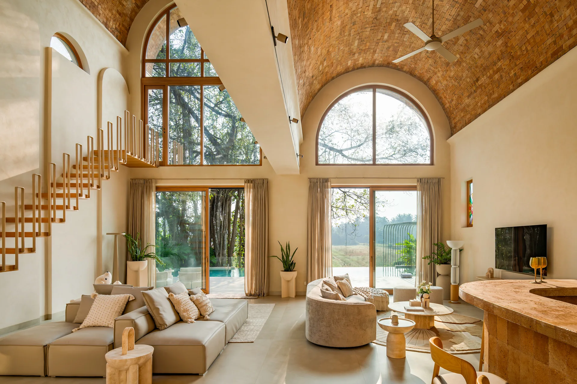 A double-height living room features a vaulted brick ceiling, large arched windows overlooking a pool, beige modular sofas, and a floating staircase.