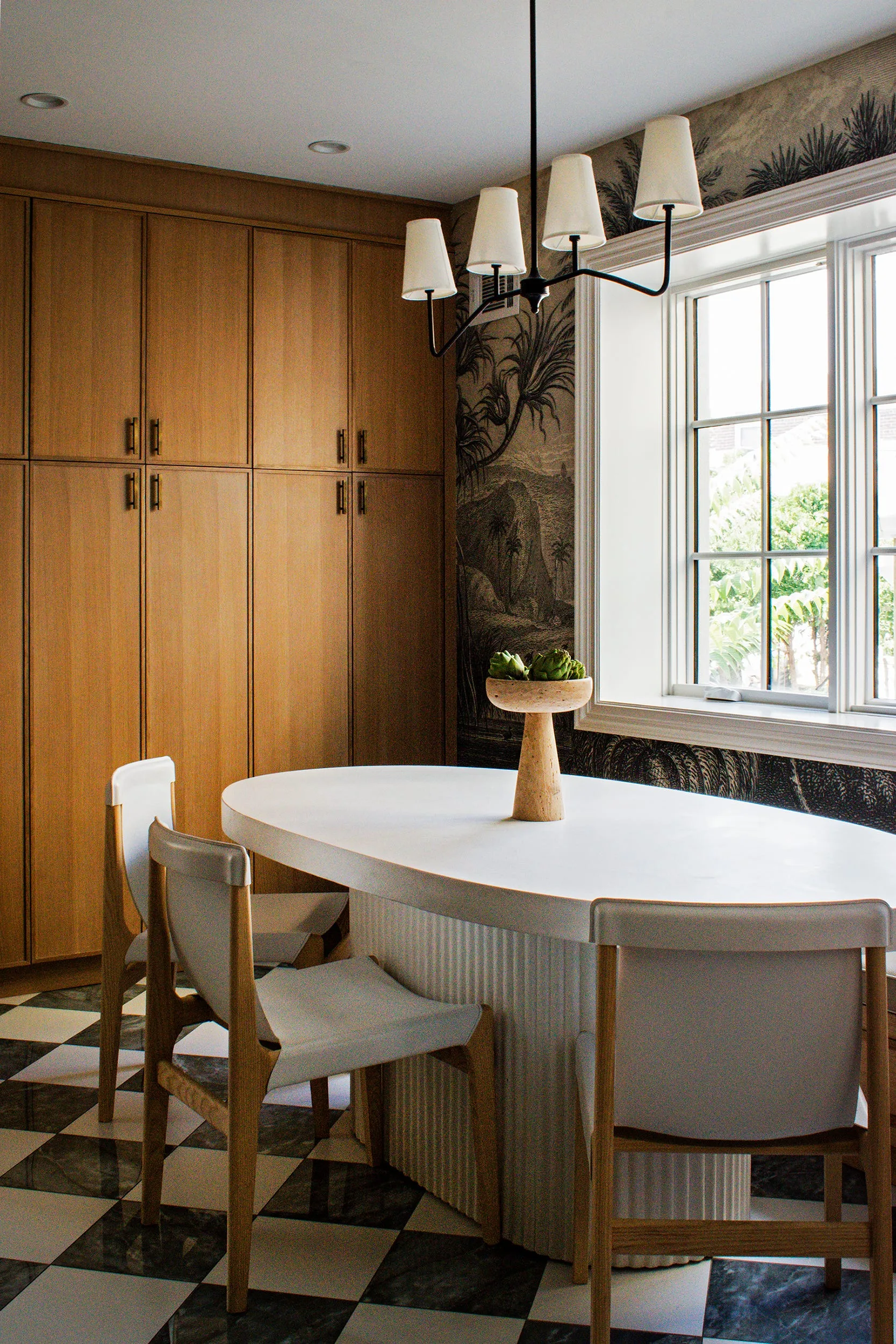 A dining room featuring a white oval table with a fluted base, light wood chairs, checkerboard floors, and scenic mural wallpaper.