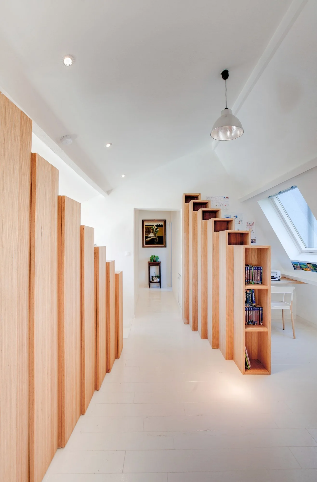 A bright hallway features custom stepped wood partitions and a matching open bookshelf beside a workspace under a skylight.