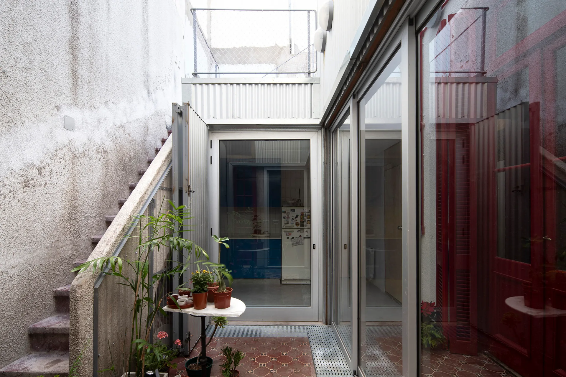 A narrow outdoor courtyard featuring a geometric tile floor, concrete stairs on the left, and sliding glass doors leading indoors.