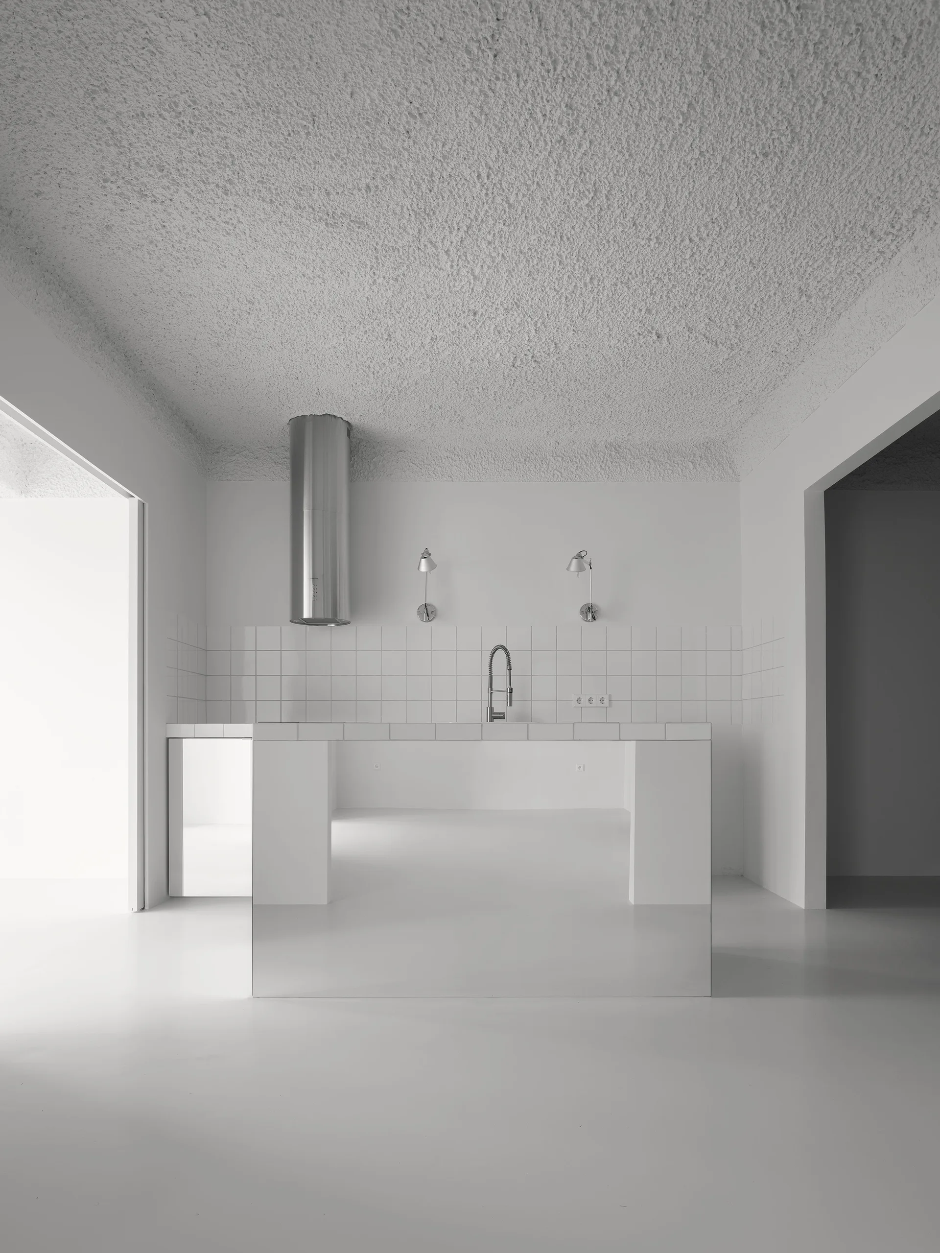 A minimalist white kitchen featuring a mirrored island base, a square tile backsplash, a cylindrical silver range hood, and a textured plaster ceiling.