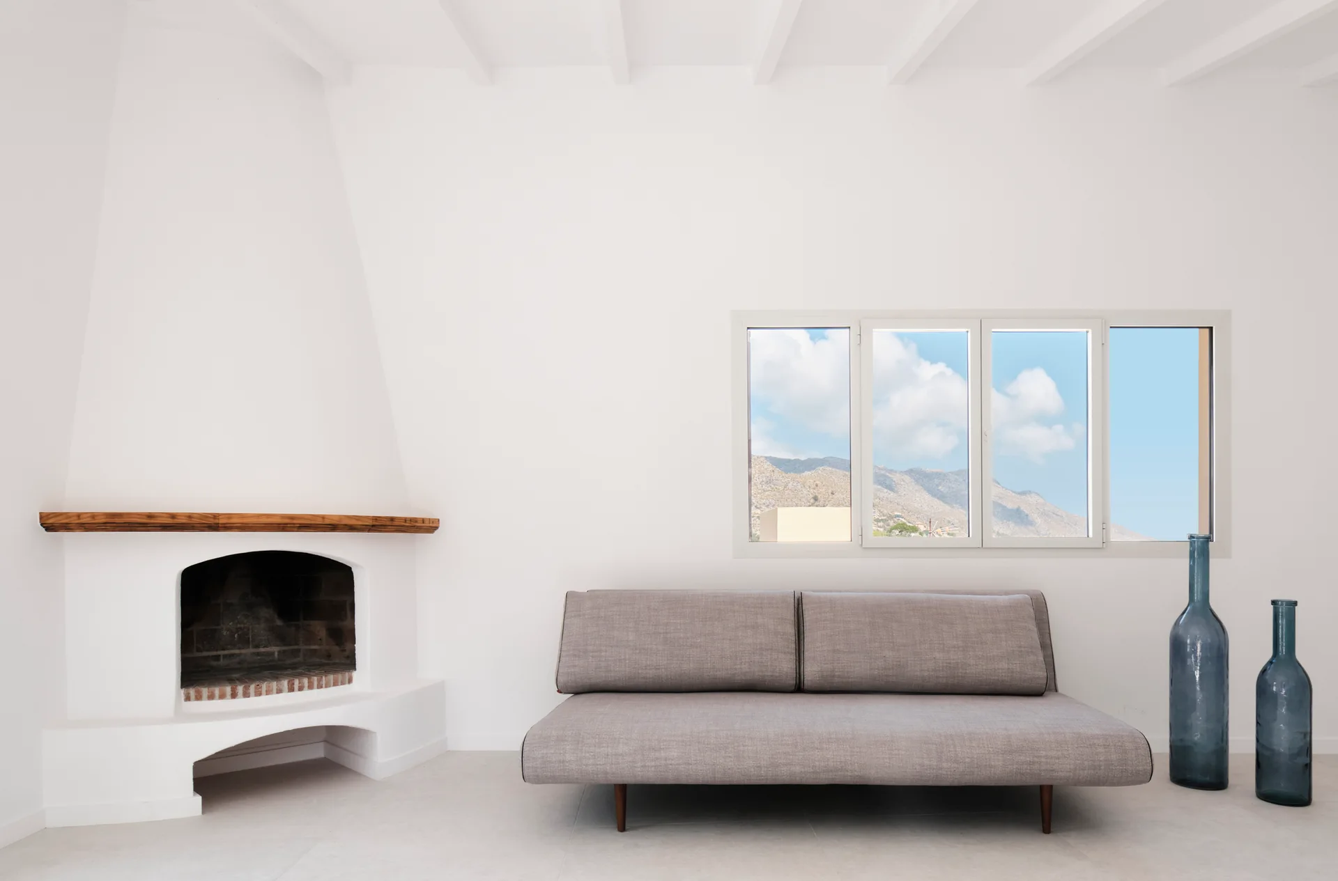 A minimalist white living room featuring a grey sofa, a plaster fireplace with a wood mantel, and tall blue glass vases.