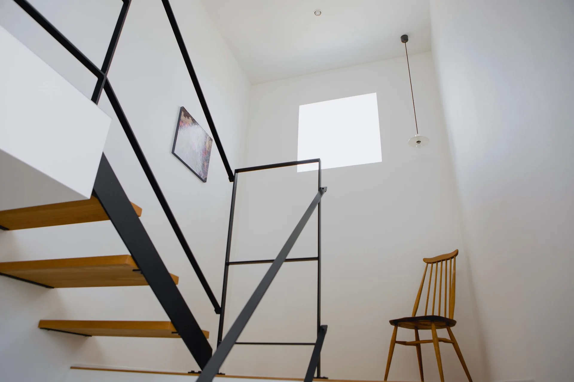 A minimalist stairwell featuring a floating wooden staircase with black metal railings, a square window, and a wooden spindle chair.