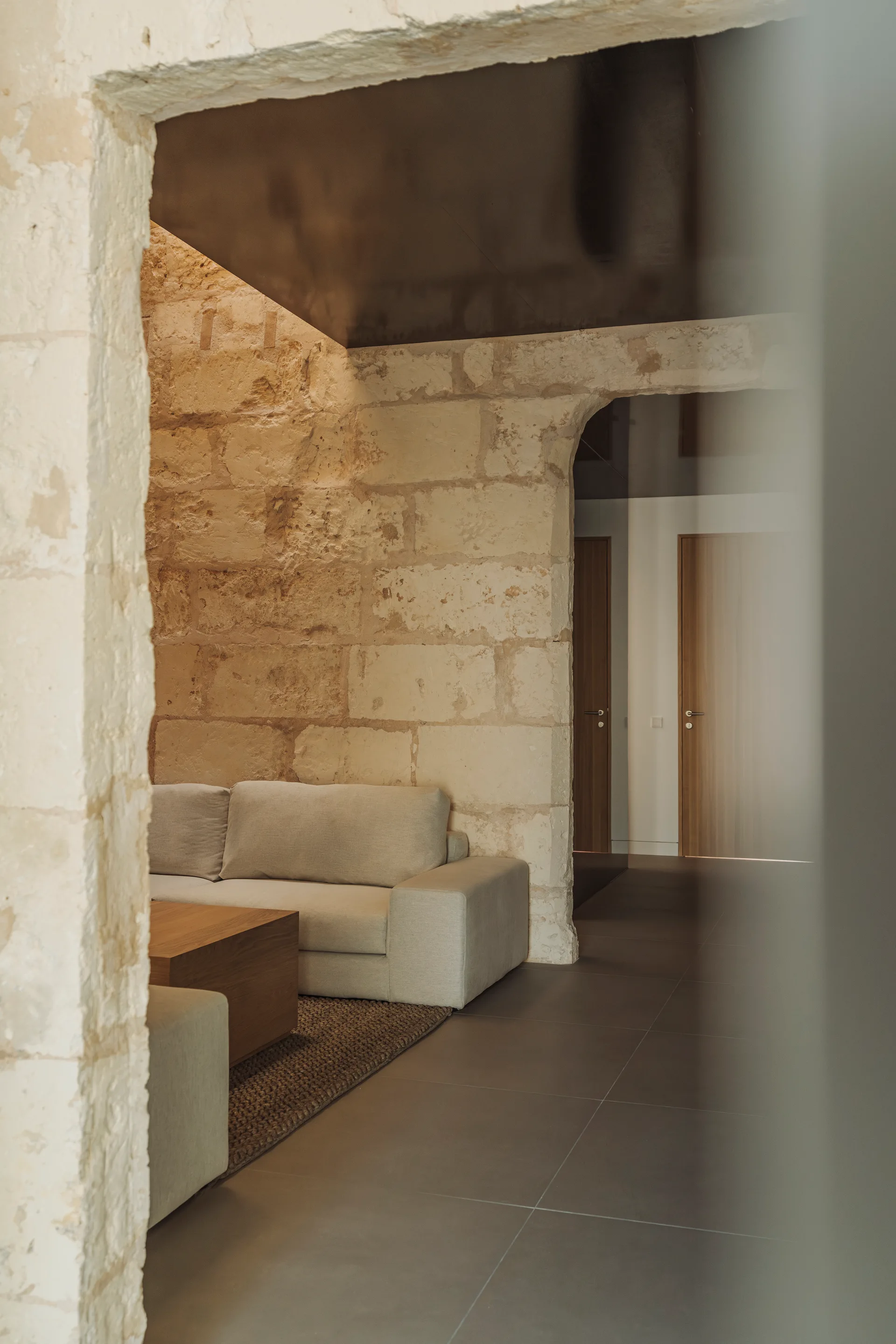 A modern living room viewed through a stone archway features a light beige sofa, a low wooden coffee table, and rough exposed masonry walls.