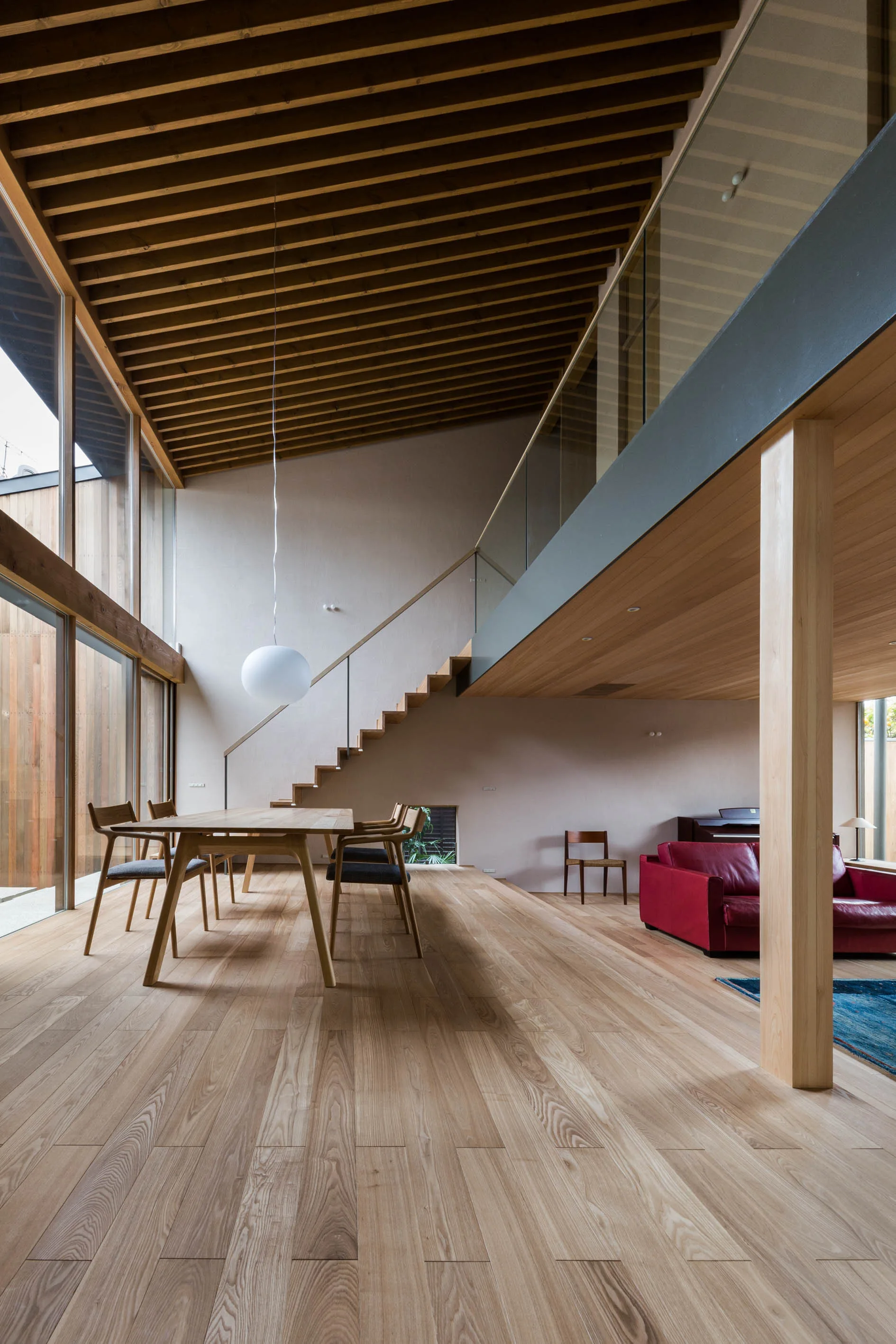 An open-plan home featuring a wooden dining set, a red sofa, and an exposed timber rafter ceiling beneath a glass-railed mezzanine.