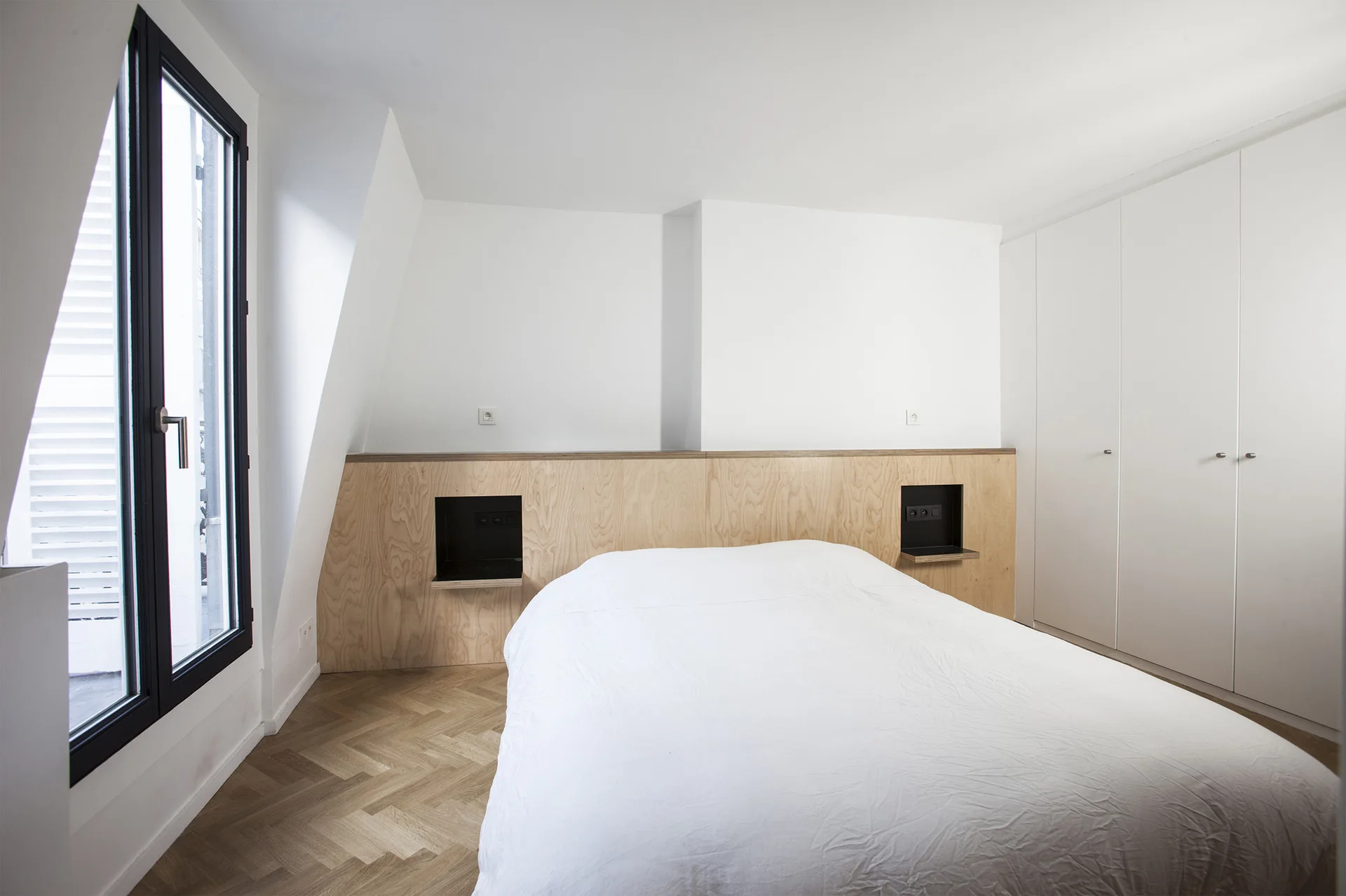A minimalist bedroom featuring a low light wood headboard with integrated black niches, white bedding, and light oak herringbone flooring.