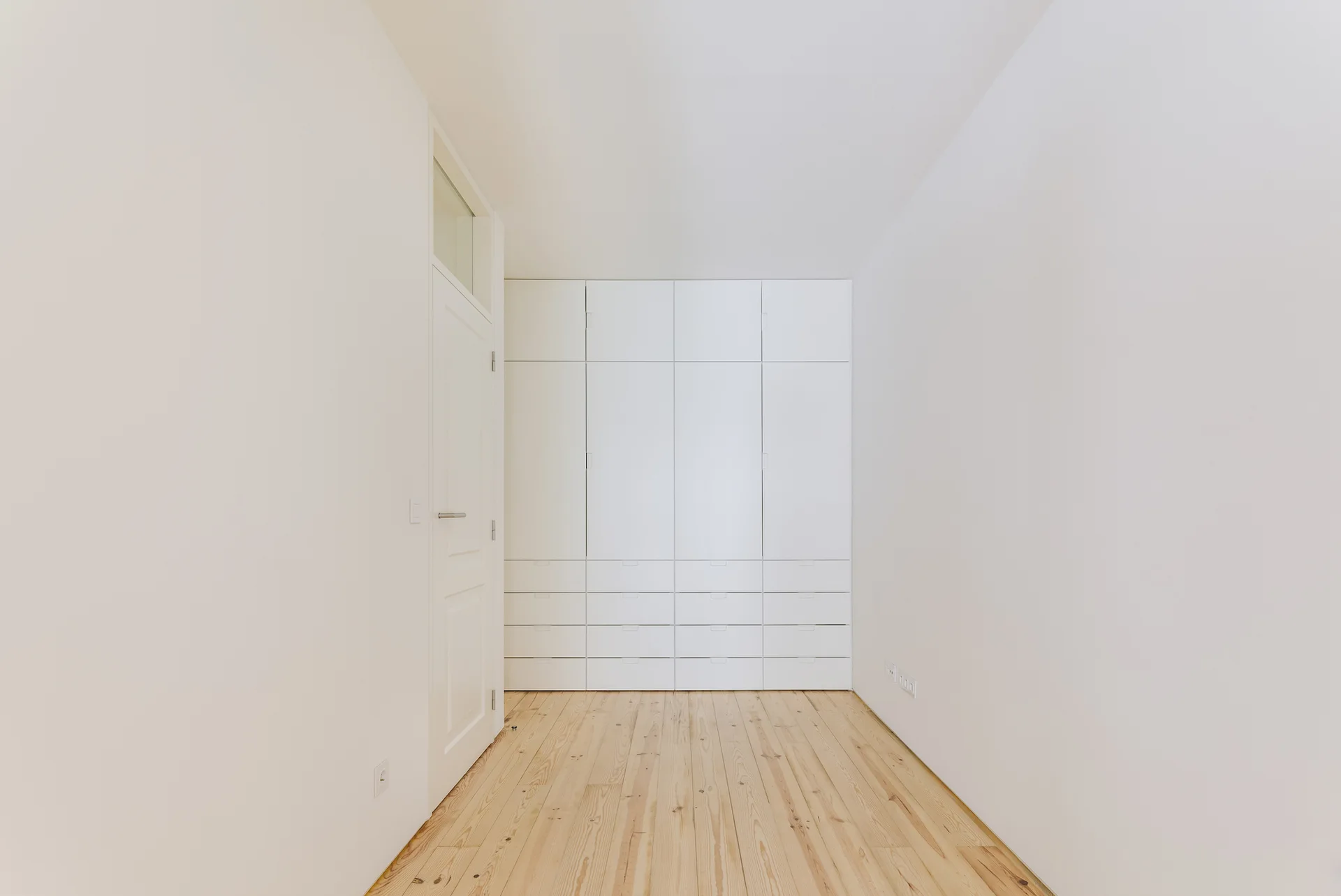 A minimalist white hallway features light pine wood flooring, a paneled door with a transom window, and a built-in floor-to-ceiling wardrobe.