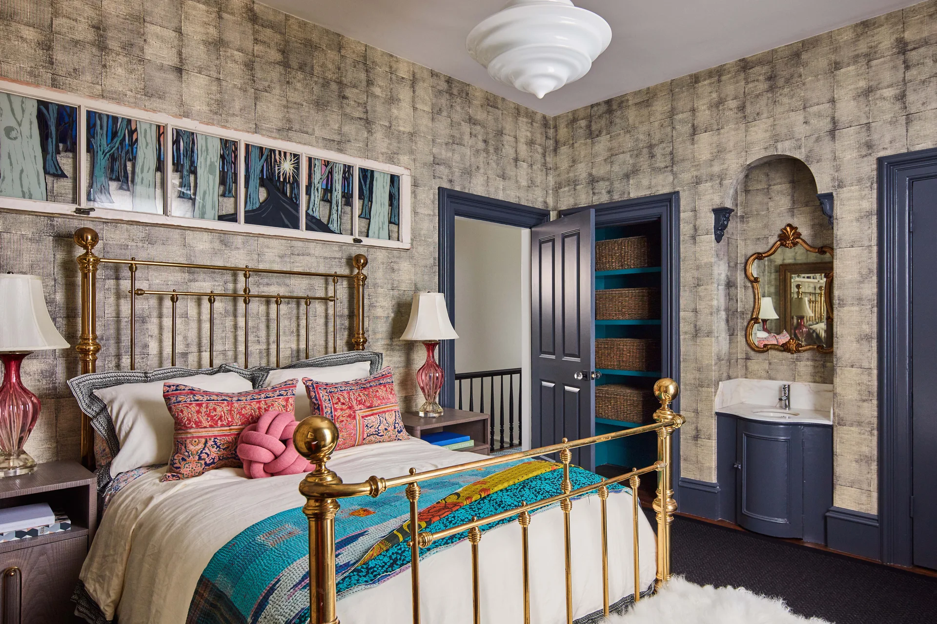 An eclectic bedroom features a brass bed frame, textured grid wallpaper, dark blue trim, and a small vanity nestled inside an arched alcove.
