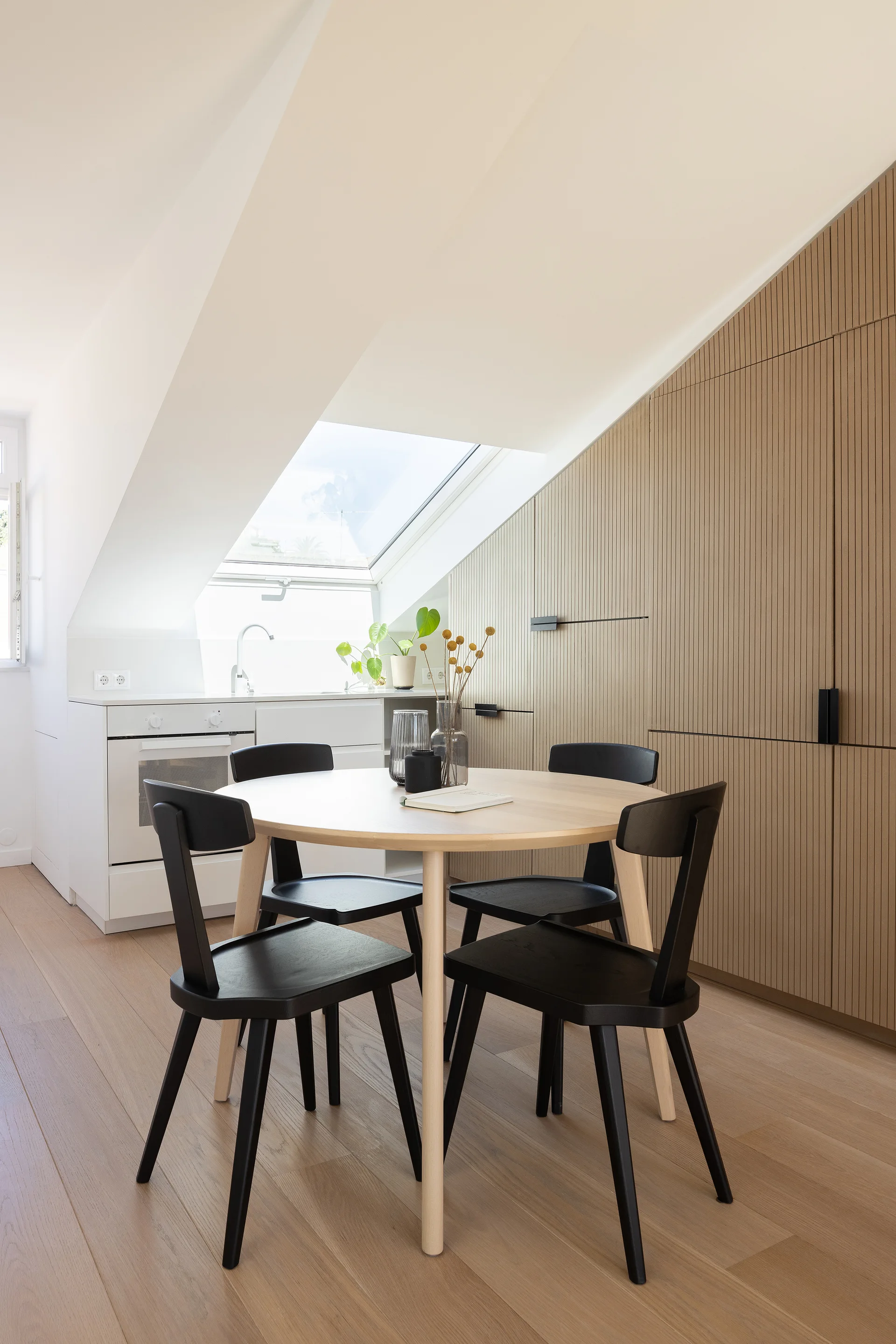 A round wooden dining table with black chairs sits in an attic kitchen featuring a slanted ceiling with a skylight and tall ribbed wood cabinets.