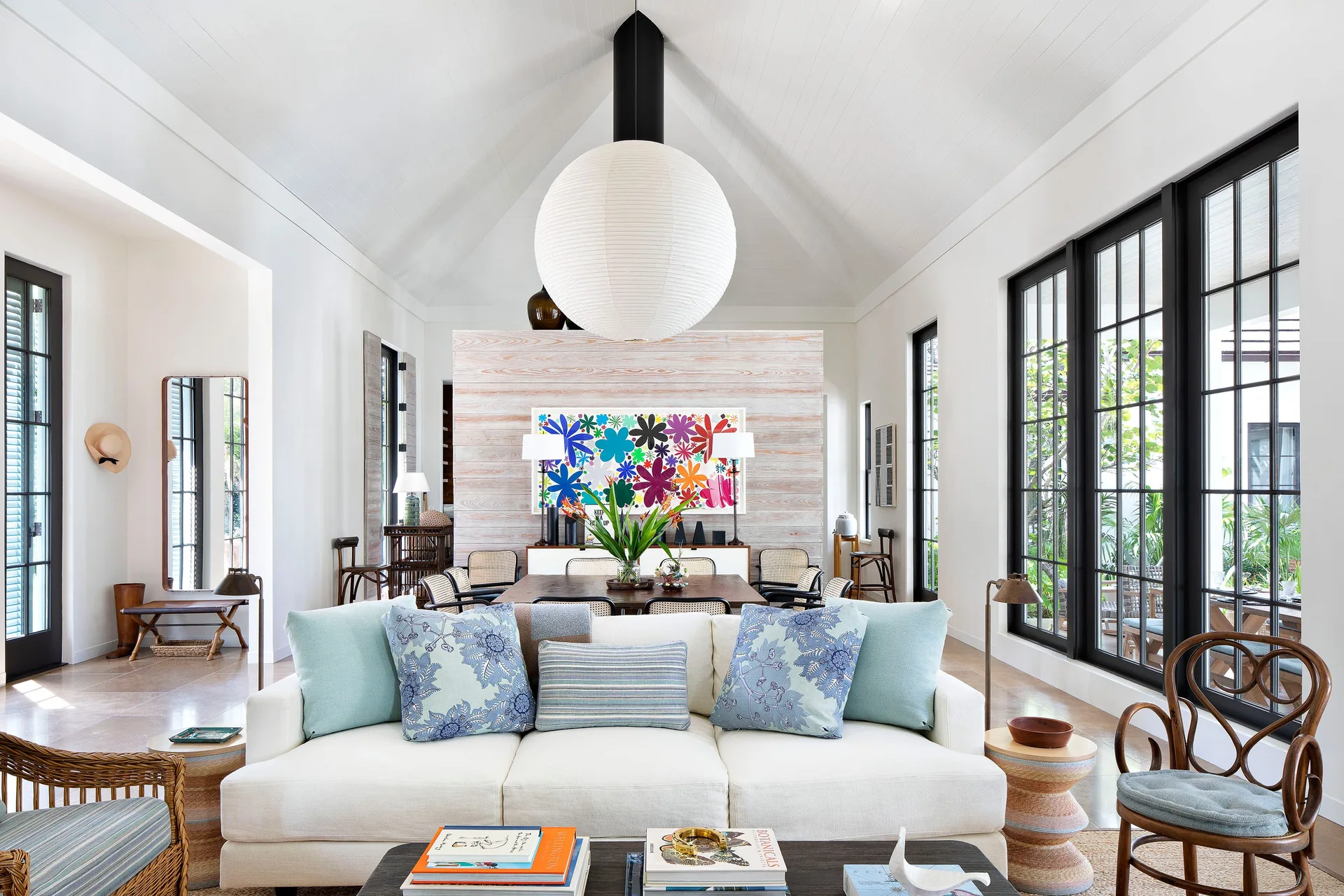 A bright living room with a white sofa, patterned blue pillows, a large paper globe pendant, and a vaulted ceiling leading to a dining area.