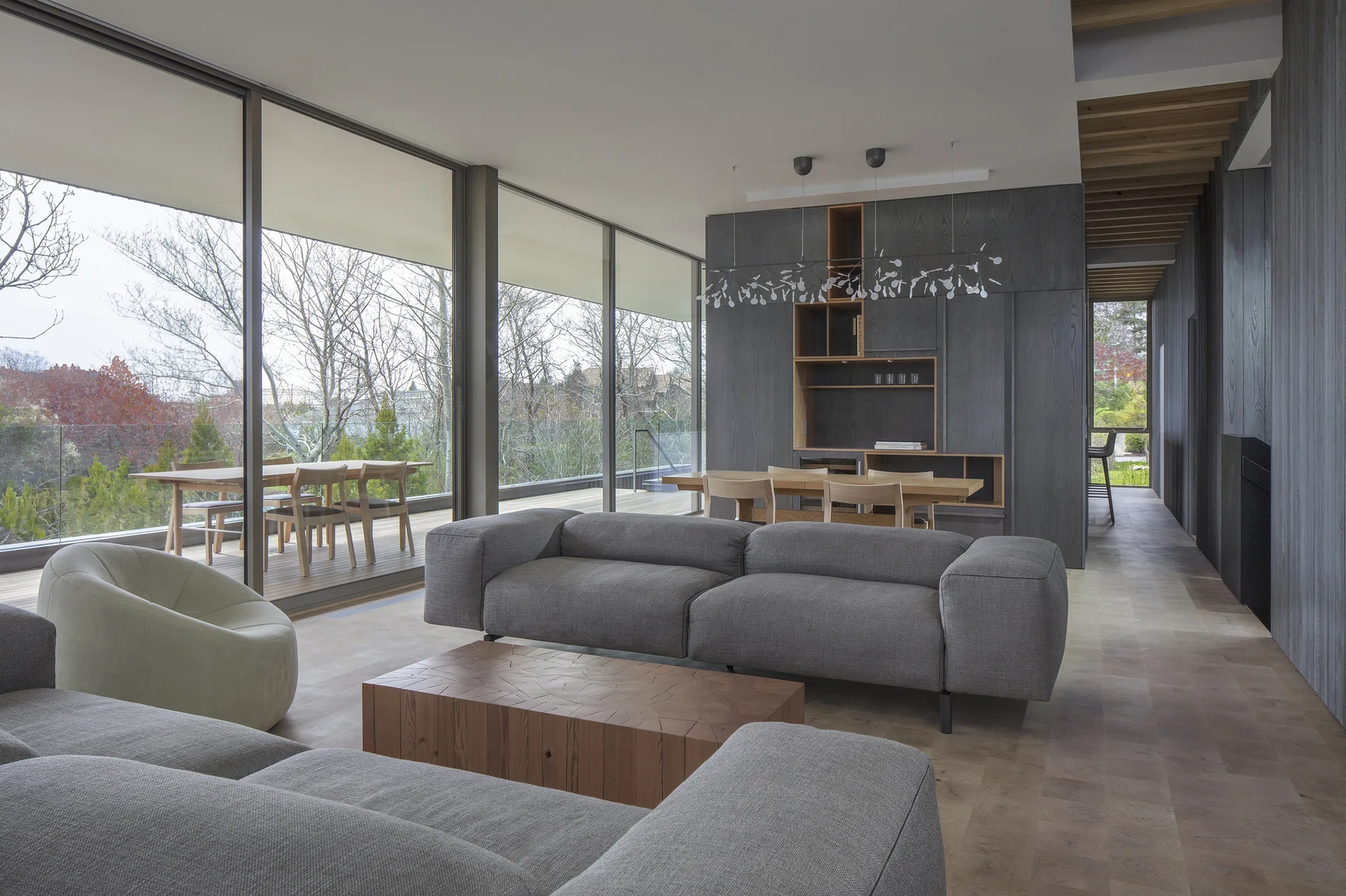 A modern open-plan living room features grey sectional sofas, a block wood coffee table, and large glass walls looking out to a wooden patio.