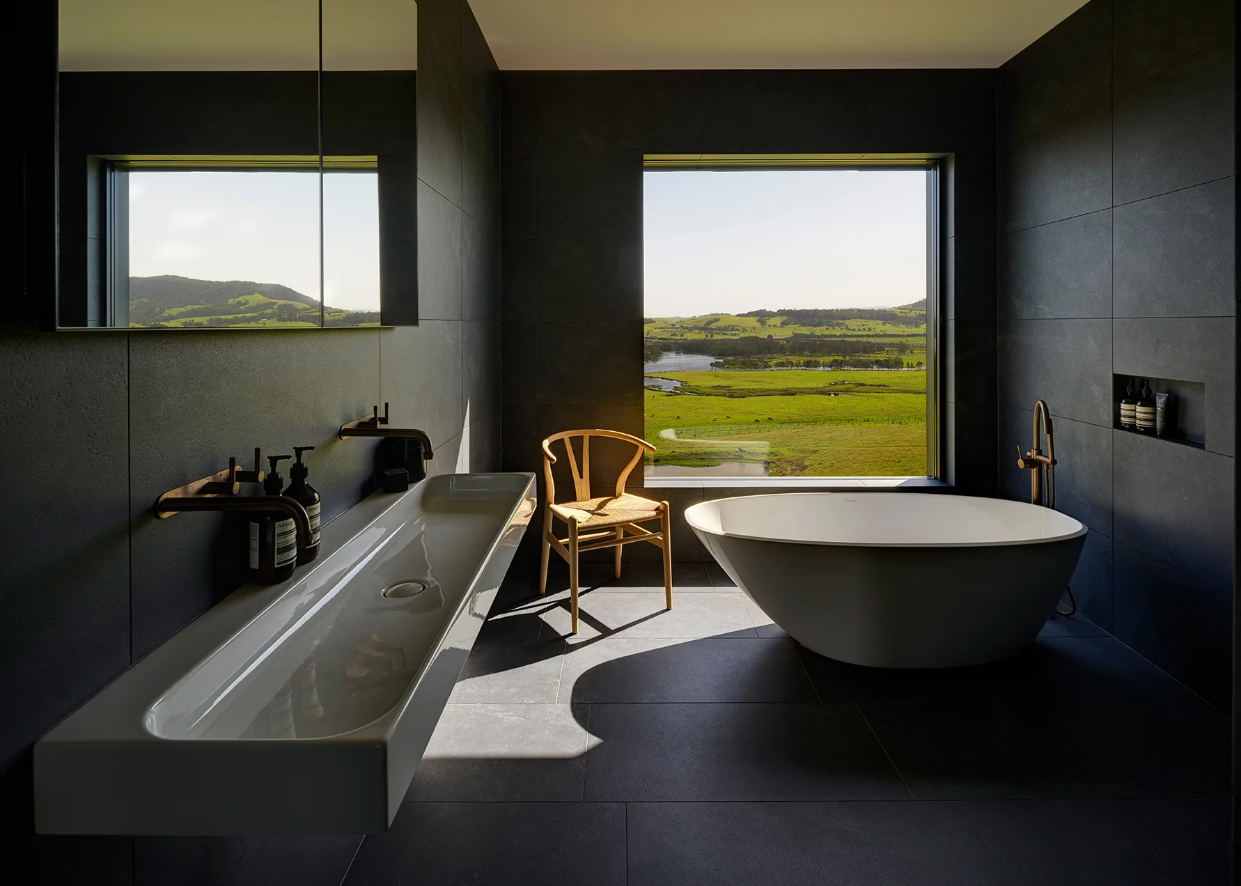 A modern bathroom features dark charcoal tiled walls and floor, a white freestanding tub, a long trough sink, and a window displaying a green landscape.