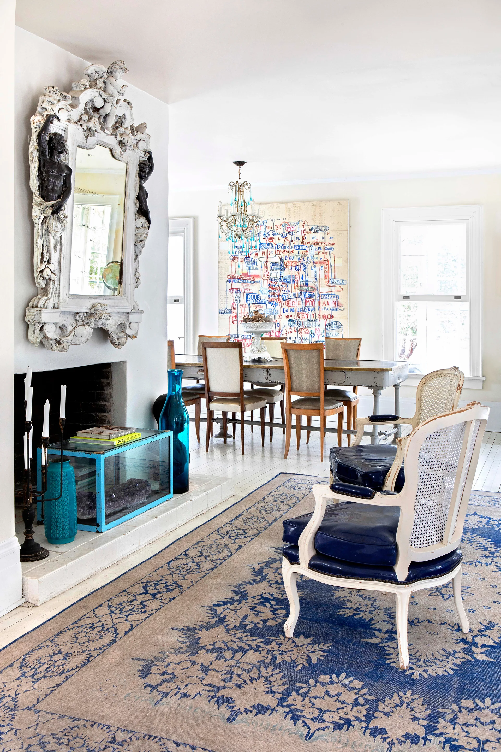 A bright dining room features a long grey table, an ornate white carved mirror over a fireplace, and blue leather armchairs on a patterned rug.