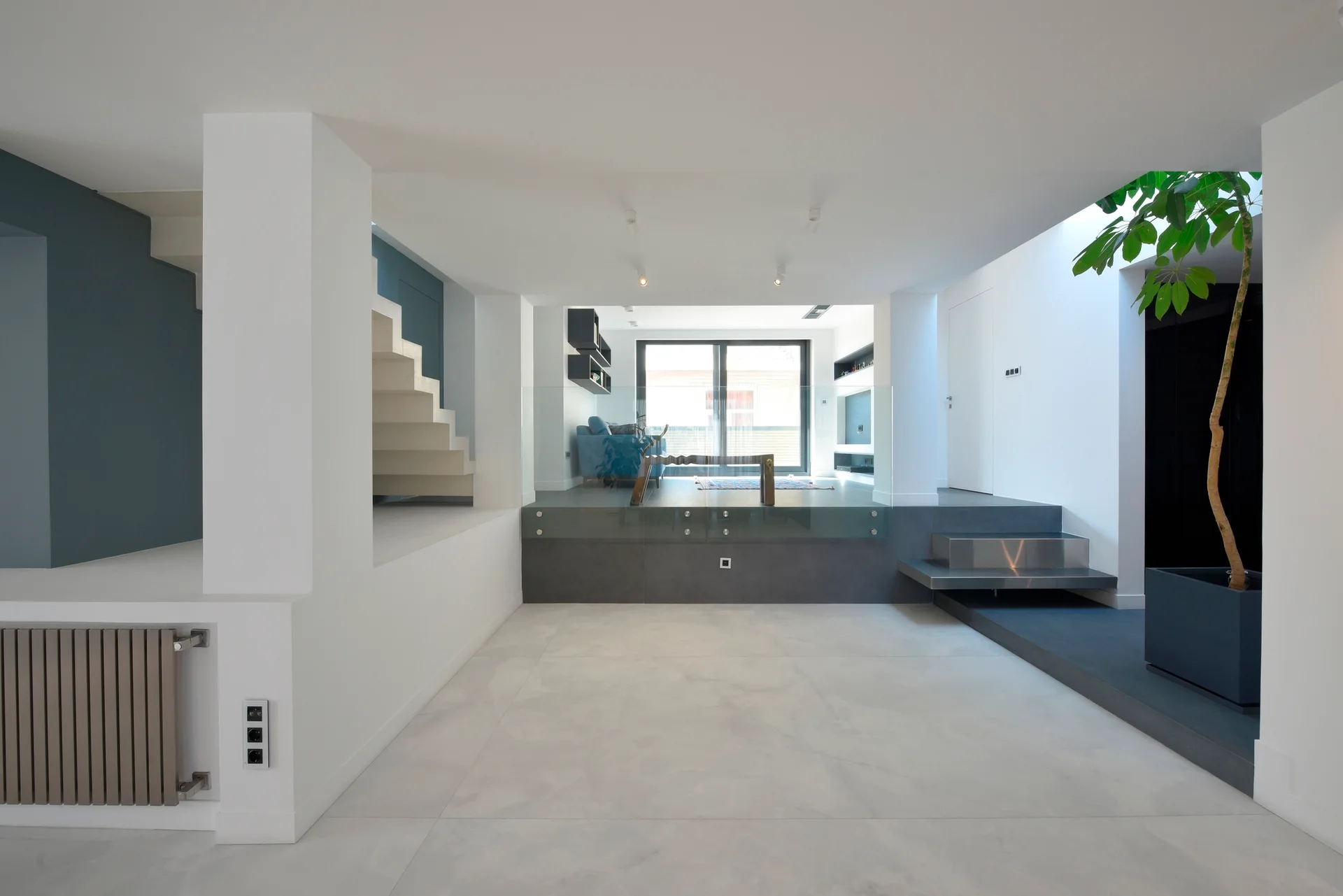 A minimalist split-level living room with large white floor tiles, a floating white staircase, glass railings, and an indoor tree in a dark grey planter.