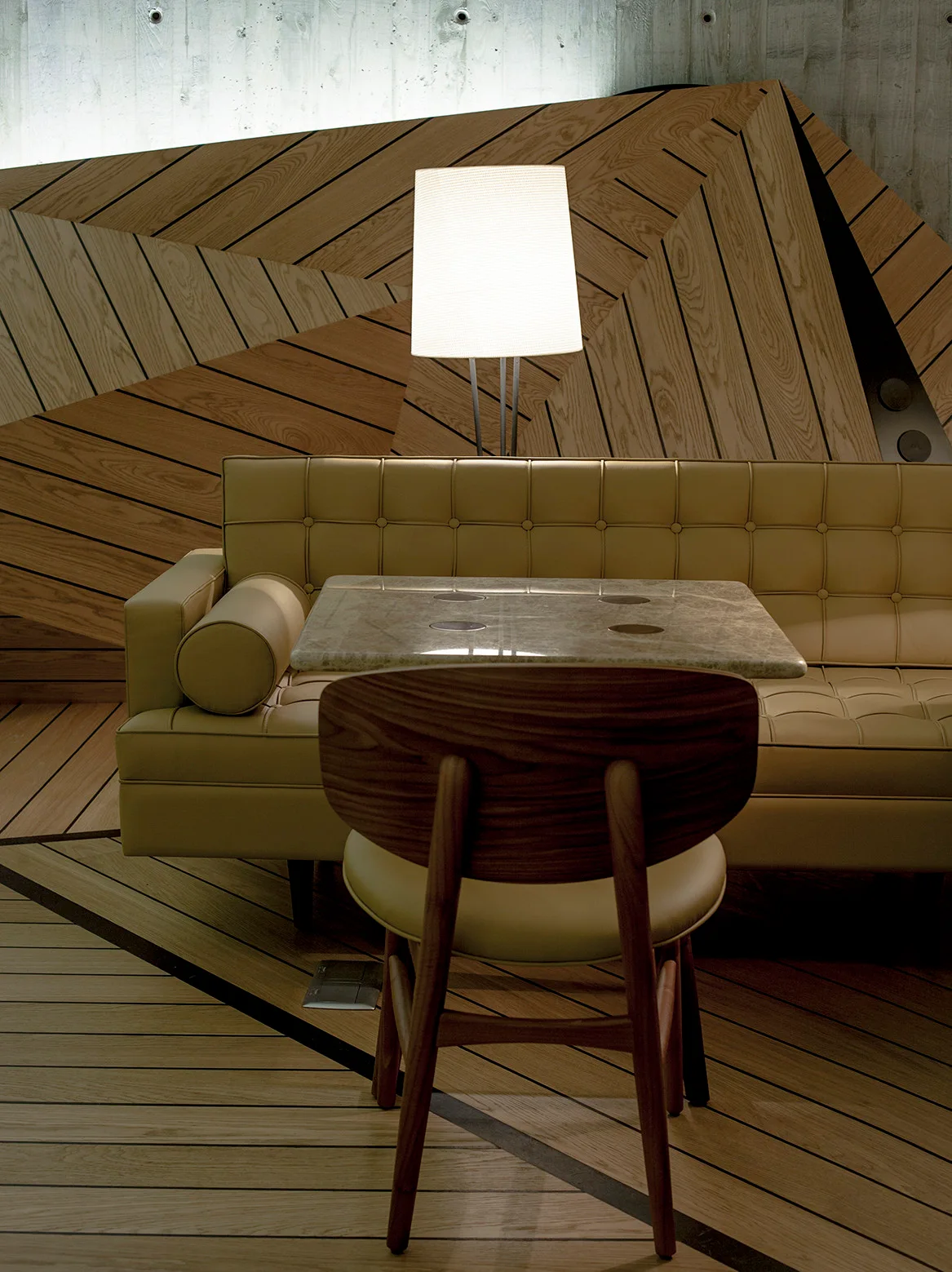 A seating area featuring a tufted cream leather sofa, a wooden chair, a stone-topped table, and a geometric wood-paneled wall illuminated by a floor lamp.
