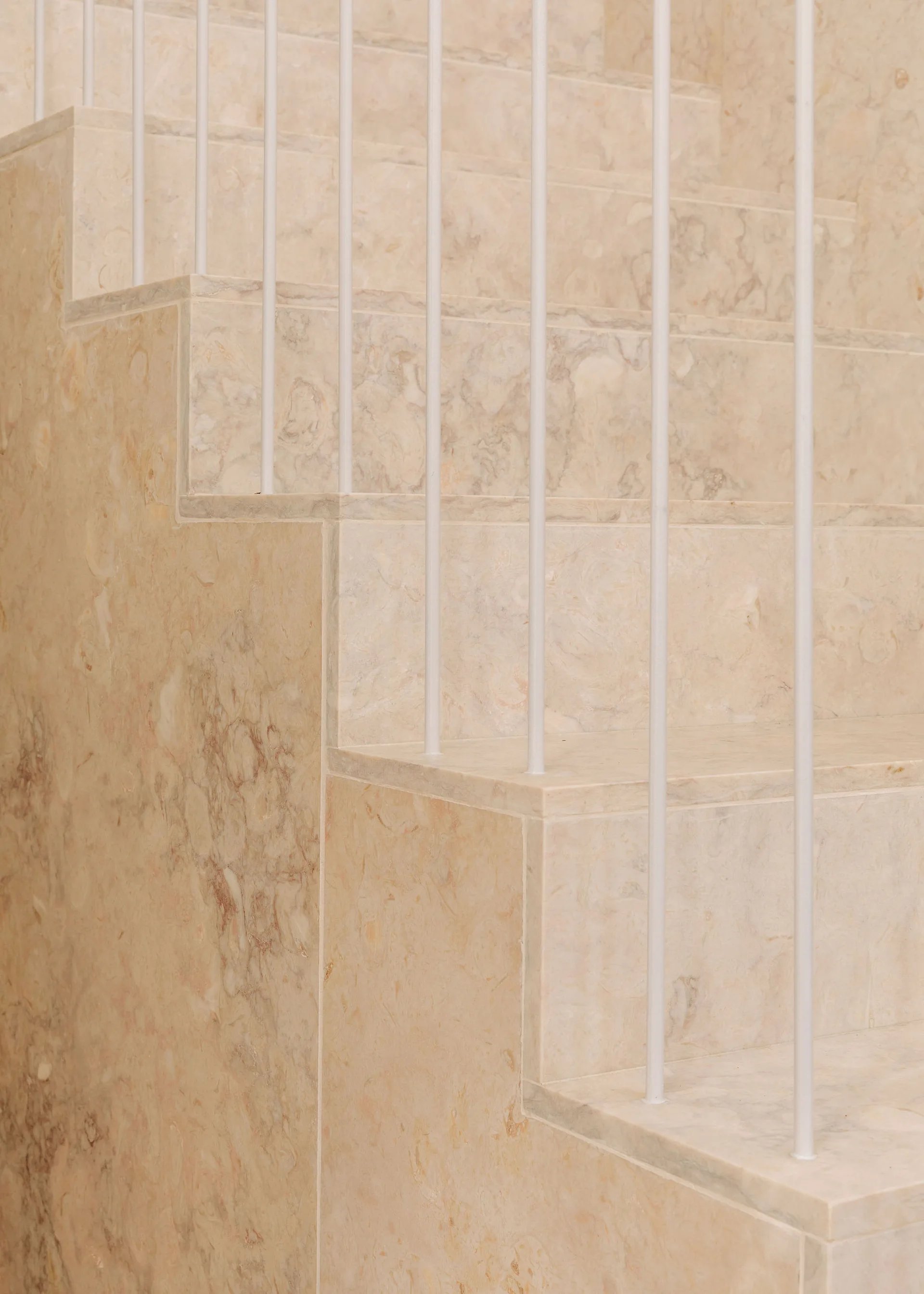 A close-up of a minimalist indoor staircase featuring polished beige marble steps and thin, vertical white metal railing spindles.