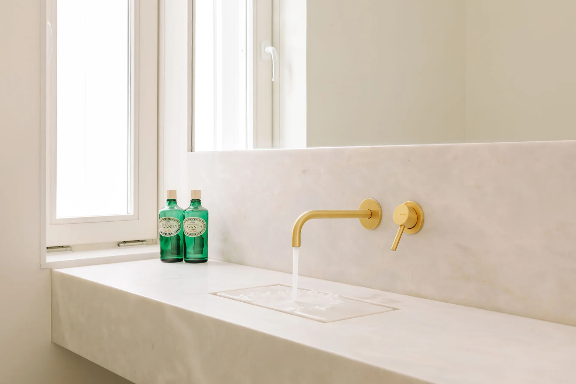 A minimalist white marble bathroom vanity with an integrated sink basin, wall-mounted brass faucet fixtures, and two green glass bottles.