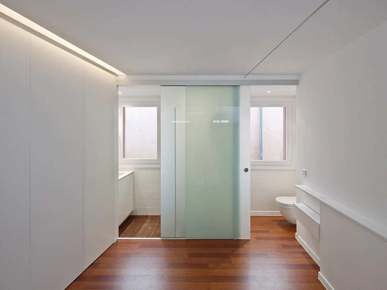 A minimalist bathroom featuring warm wood flooring, a frosted glass sliding door, seamless white closets, and two small windows.