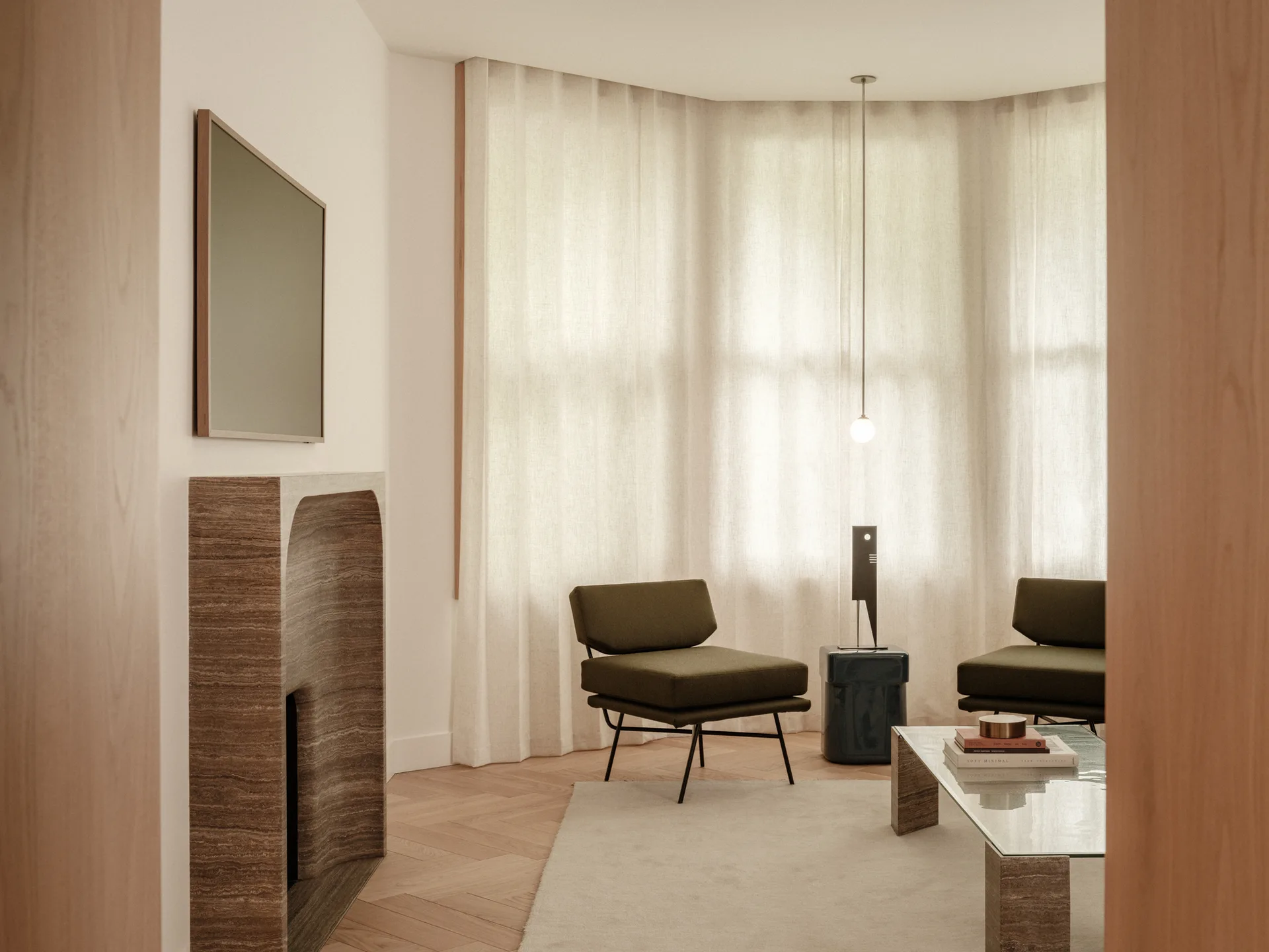 A minimalist living room features two olive green lounge chairs, a glass-topped coffee table, and an arched travertine fireplace on herringbone floors.