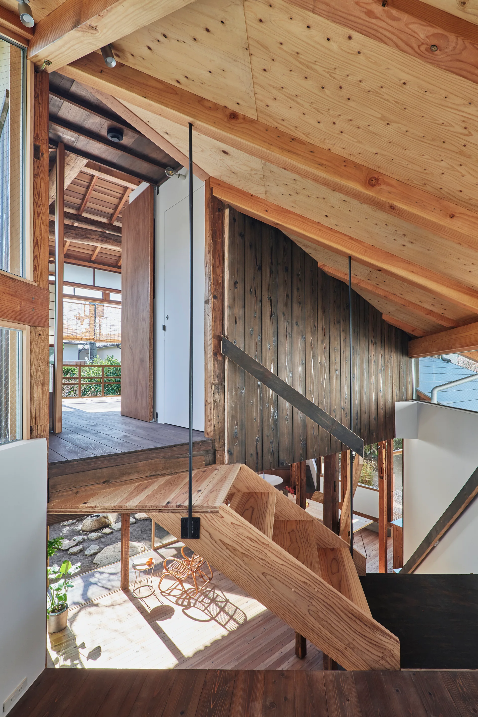 A central timber staircase connects multiple levels in a modern wood-framed house featuring exposed ceiling rafters and dark wood paneling.