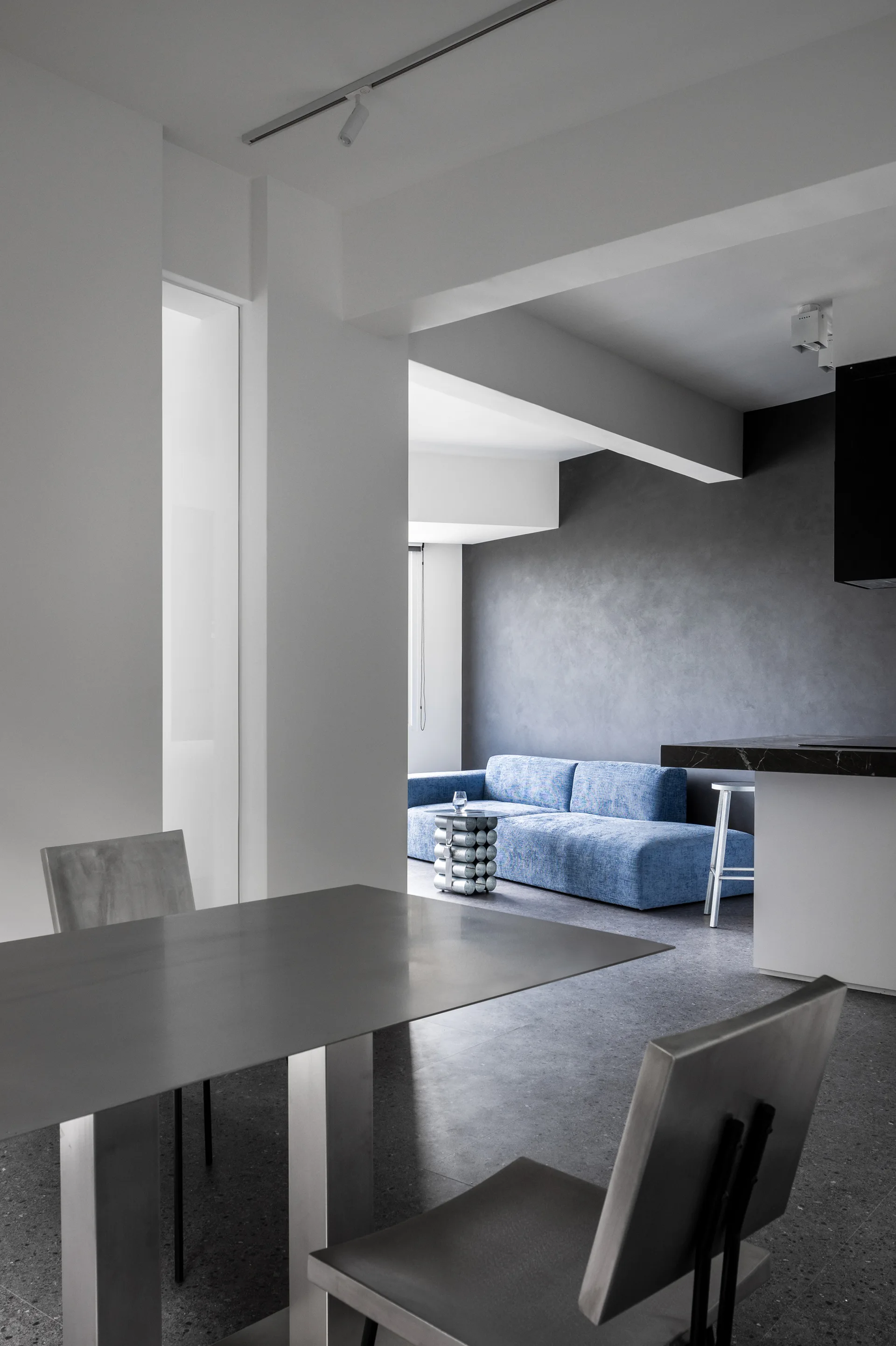 A minimalist open-concept apartment featuring a brushed metal dining table and chairs in the foreground with a textured blue sofa against a dark gray wall.
