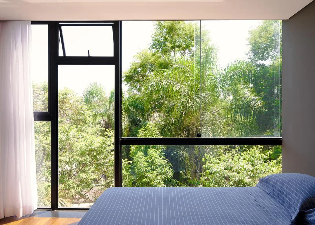 A modern bedroom features blue striped bedding next to a large black-framed window overlooking green treetops, with a sheer white curtain.