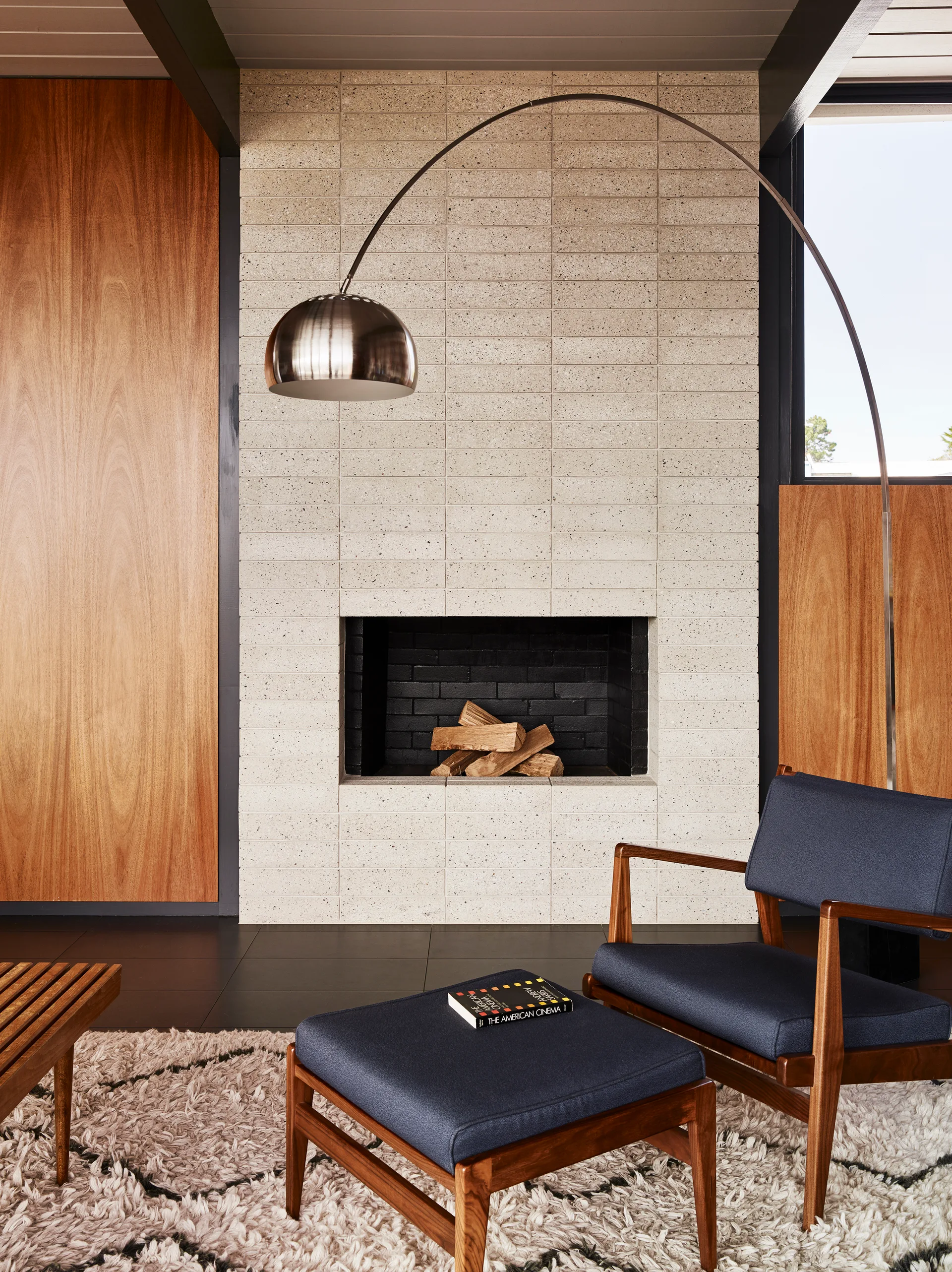 A mid-century modern living room features a light speckled tile fireplace, wood wall paneling, a blue armchair with ottoman, and a chrome arc lamp.