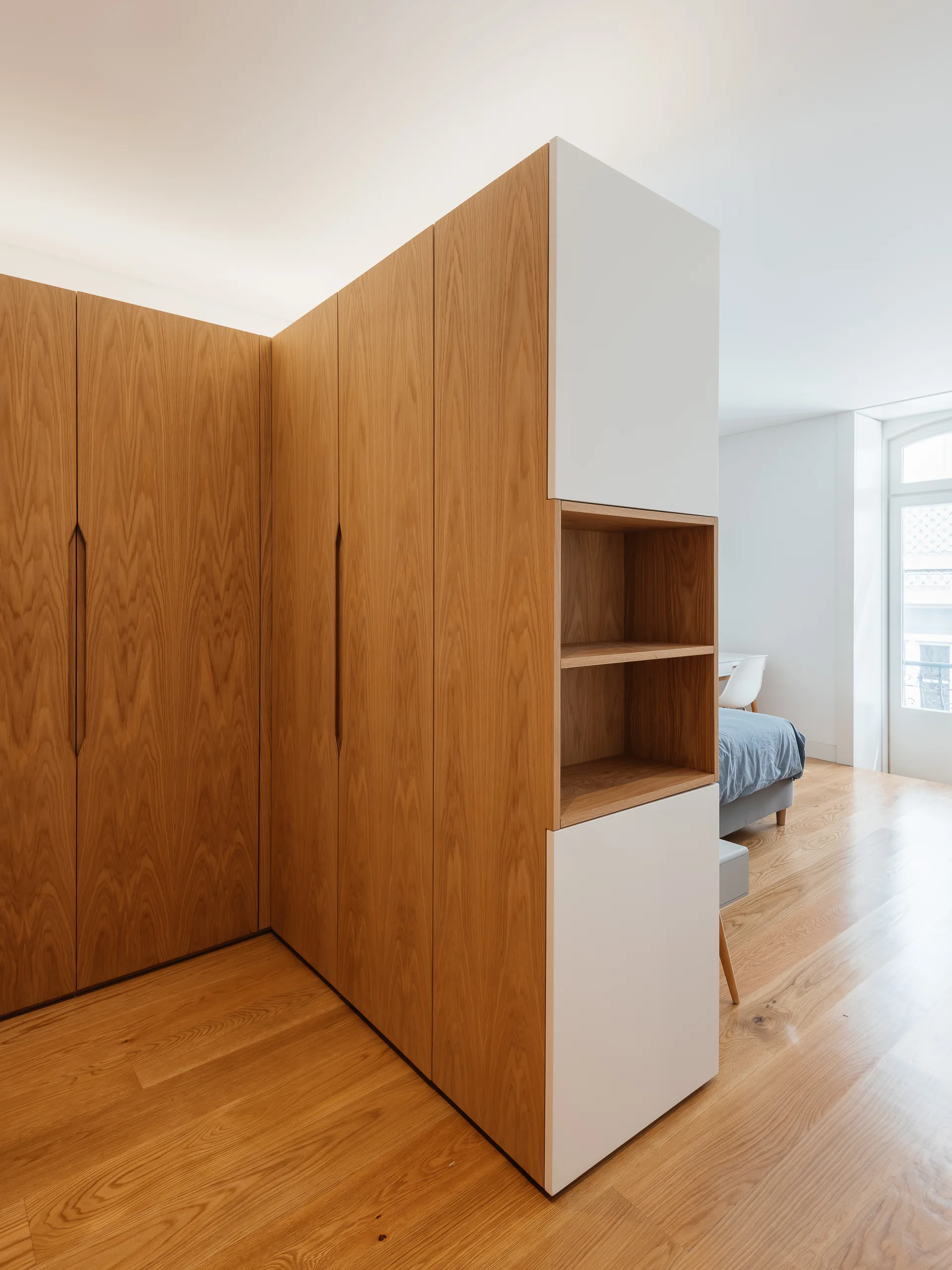 A custom L-shaped wooden wardrobe features integrated white panels and recessed open shelves on a light oak floor in a modern bedroom.