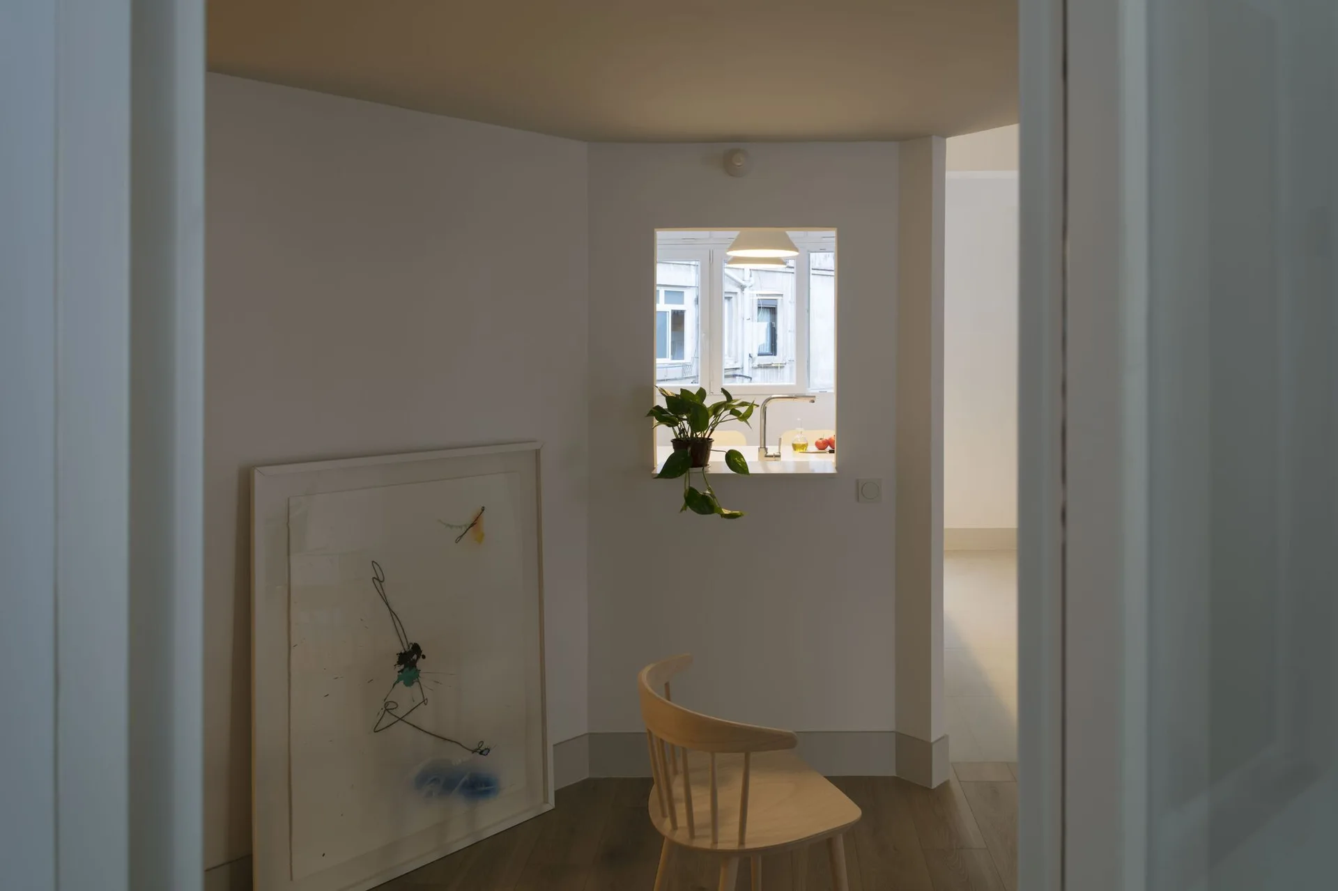 A light wood spindle chair and a large framed artwork sit in a minimalist white room next to an interior pass-through window with a plant.