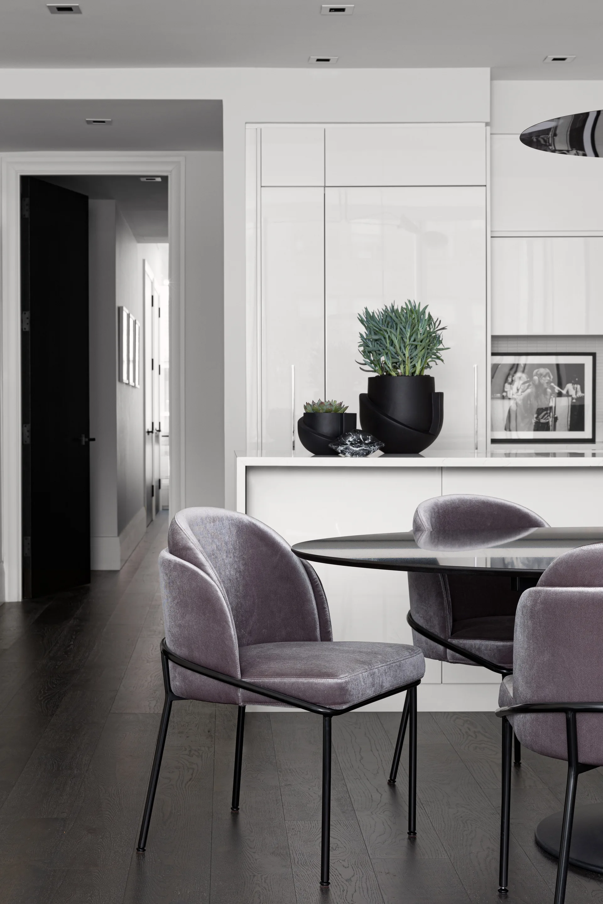 A modern dining area features grey velvet chairs around a black circular table set against glossy white kitchen cabinets.