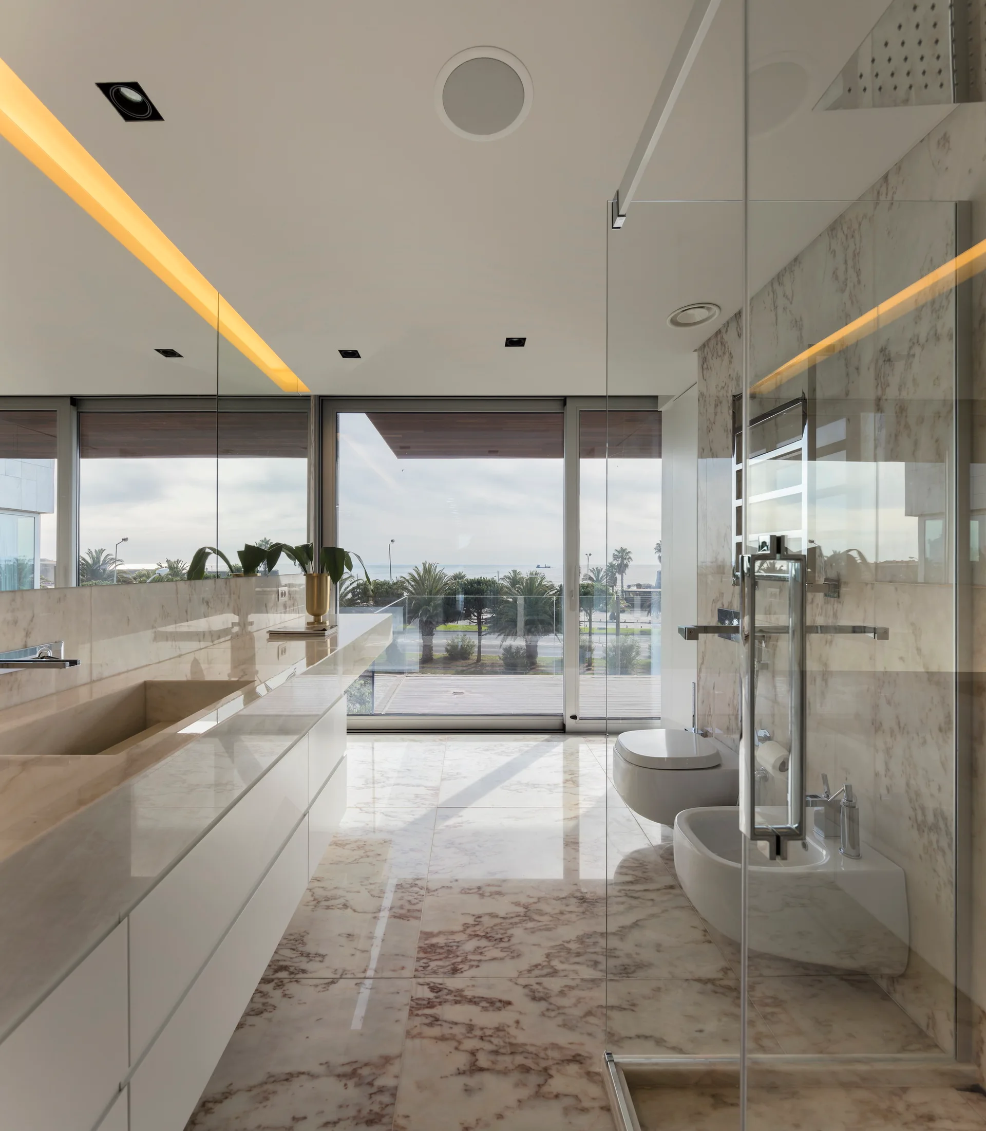 A modern bathroom features a white floating vanity, polished marble floors, a glass-walled shower, and large windows looking out to palm trees.