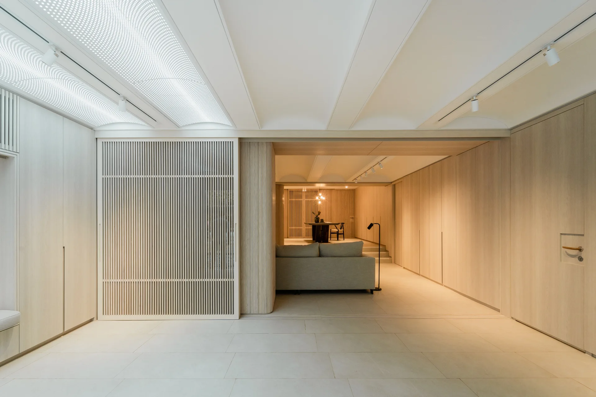 A modern, minimalist living space featuring extensive light wood wall paneling, a large slatted sliding door, and a light gray sofa.