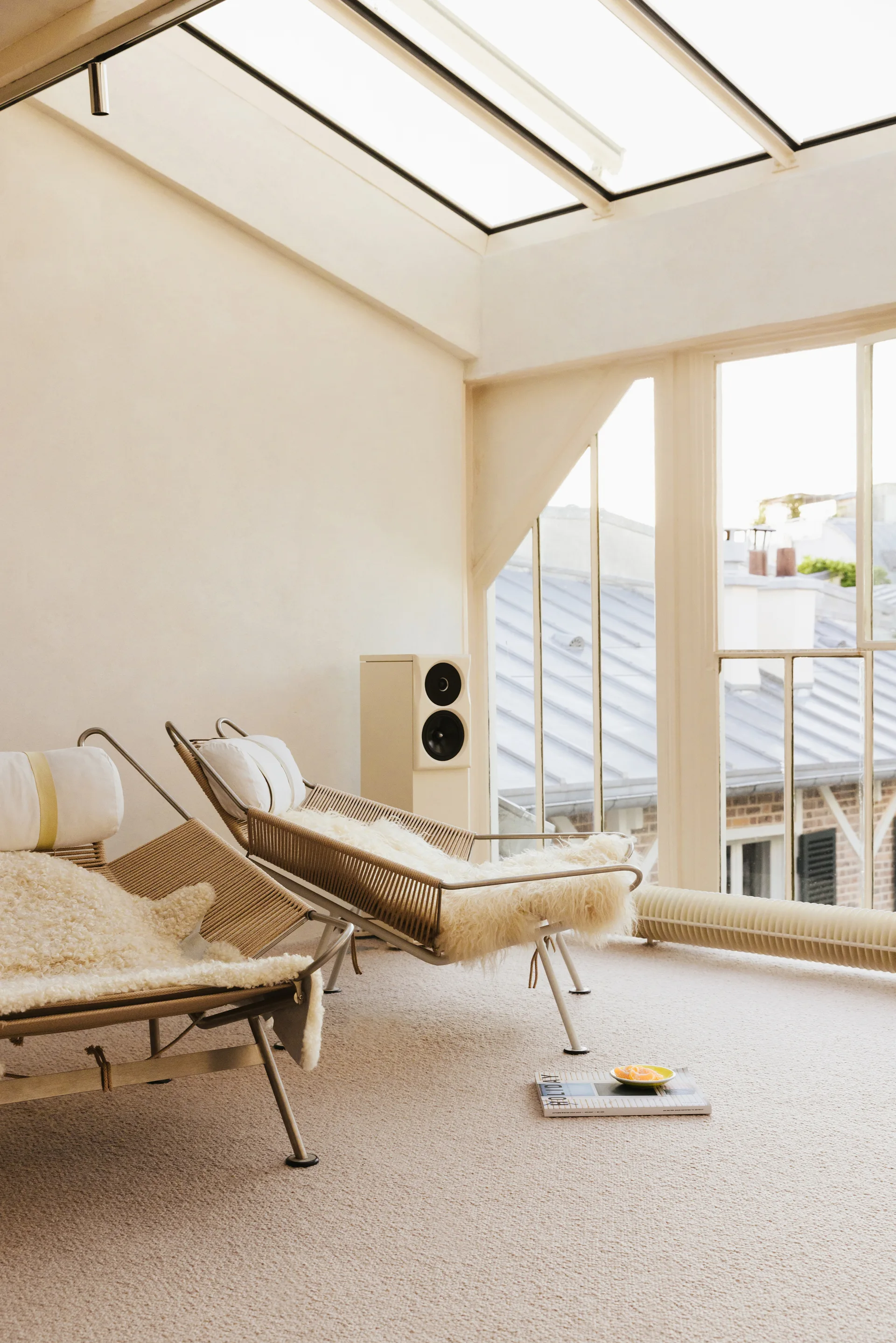 Two woven lounge chairs topped with fluffy sheepskin sit on a beige carpeted floor in a sunroom under a skylight and window.