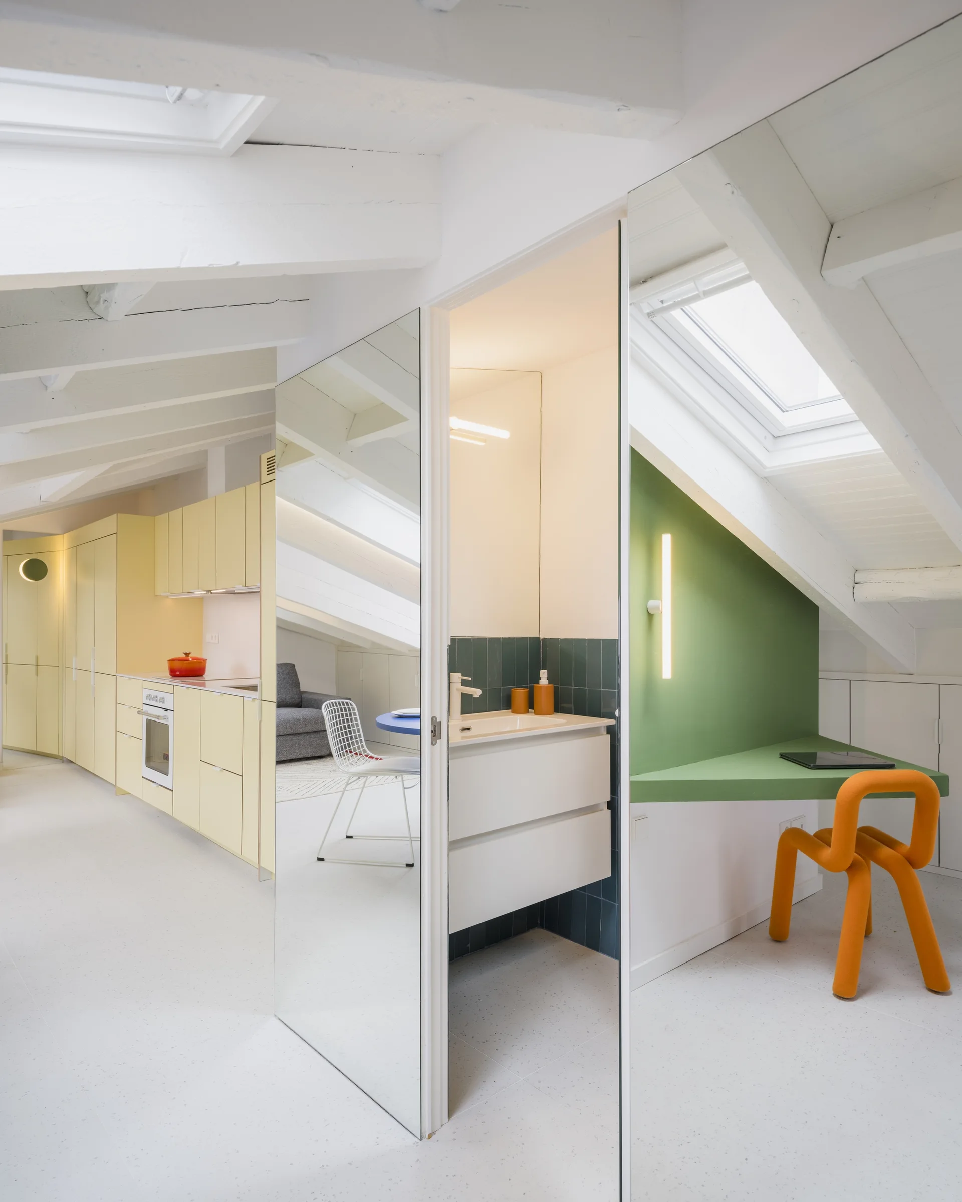 A modern loft apartment under a slanted white ceiling with a yellow kitchen, a mirrored partition, and a green built-in desk with an orange chair.