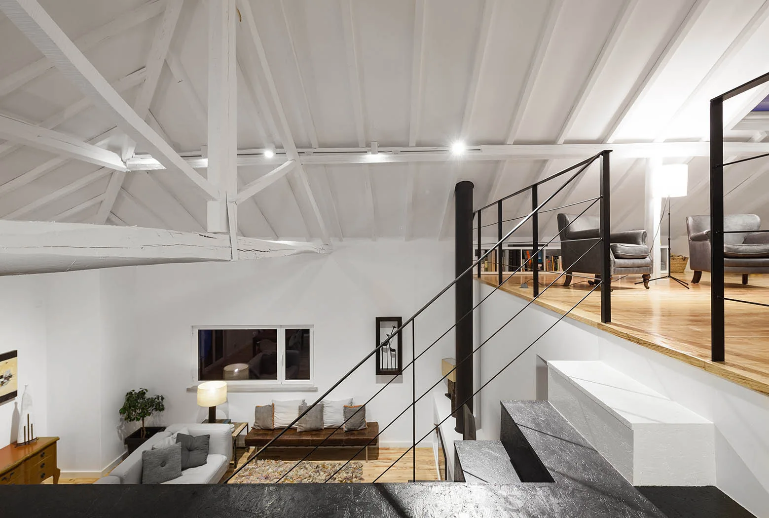 A split-level home interior featuring black metal stairs leading to a loft library under a vaulted white ceiling with exposed wooden beams.