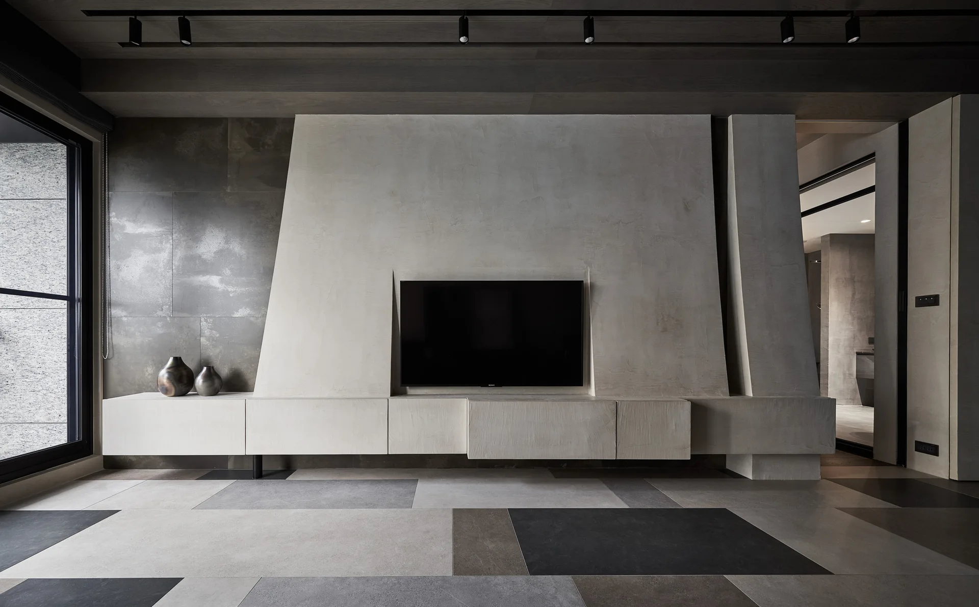 A minimalist living room featuring a textured concrete TV wall, matching low-profile built-in console, and large-format geometric floor tiles.