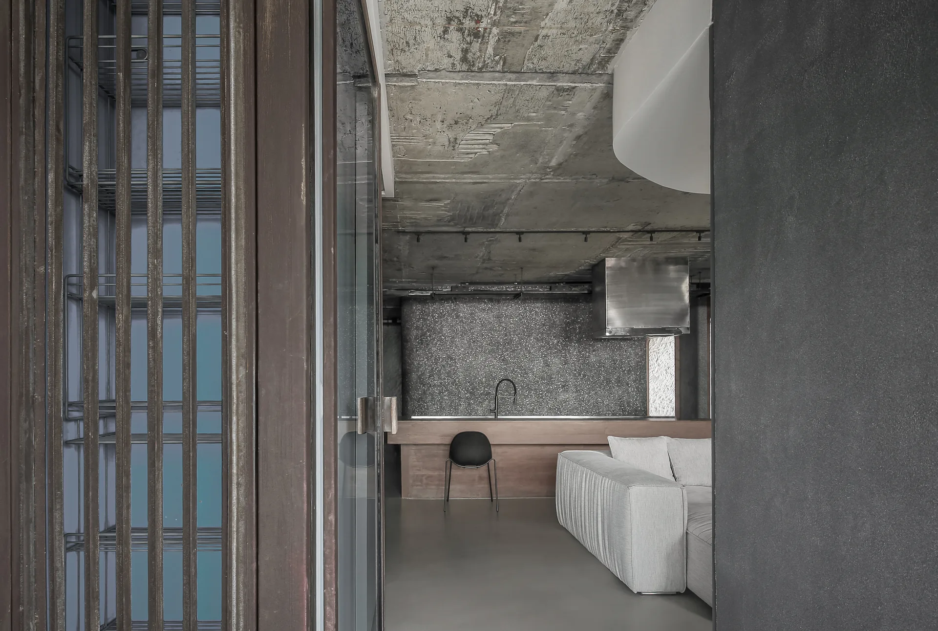An industrial living room features a raw concrete ceiling, a light gray sofa, and a wood kitchen island with a black chair.