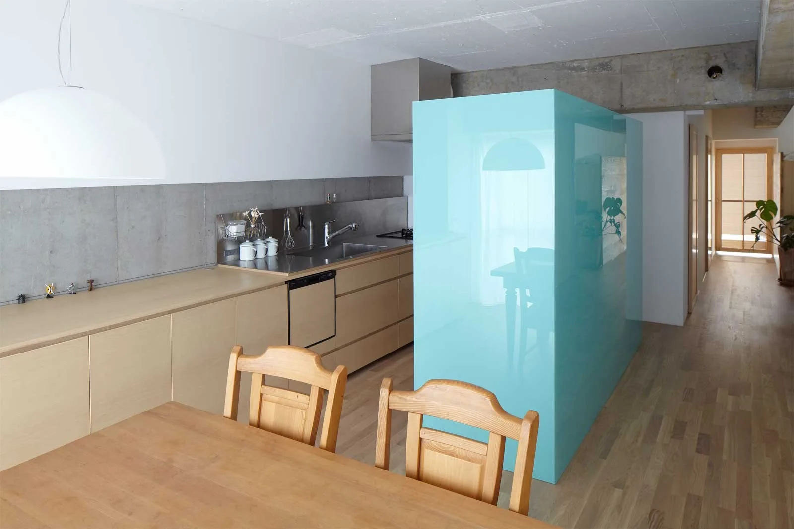 A minimalist kitchen features a glossy cyan partition wall, light wood cabinets, and an exposed concrete backsplash near a wooden dining table.