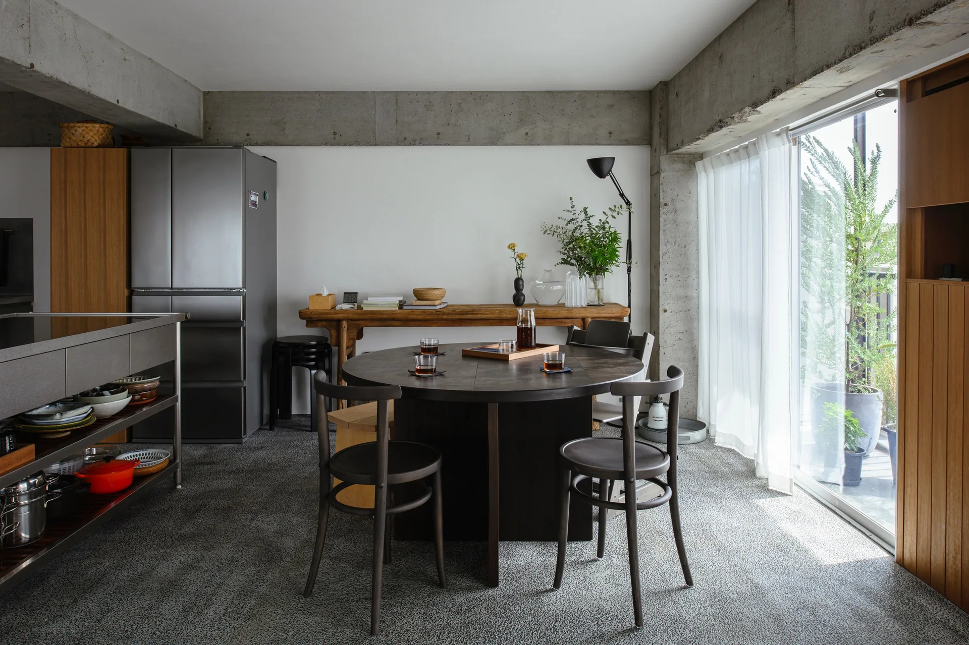 A minimalist dining room features a round dark wood table, matching chairs, a rustic console table, and exposed concrete beams next to a sliding glass door.