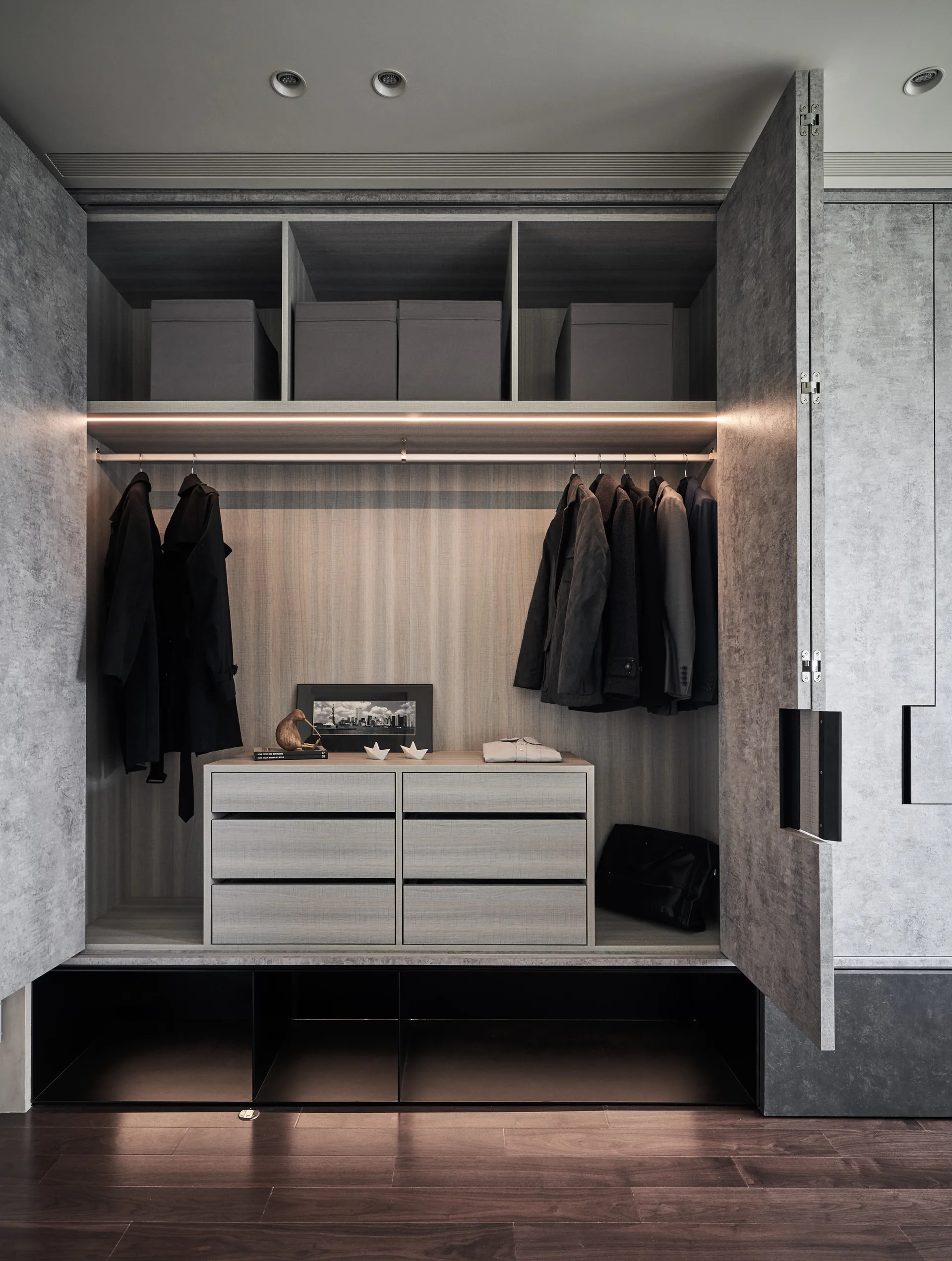 A modern wardrobe with gray concrete-effect doors open to reveal wood-grain drawers, hanging clothes, and integrated shelf lighting.