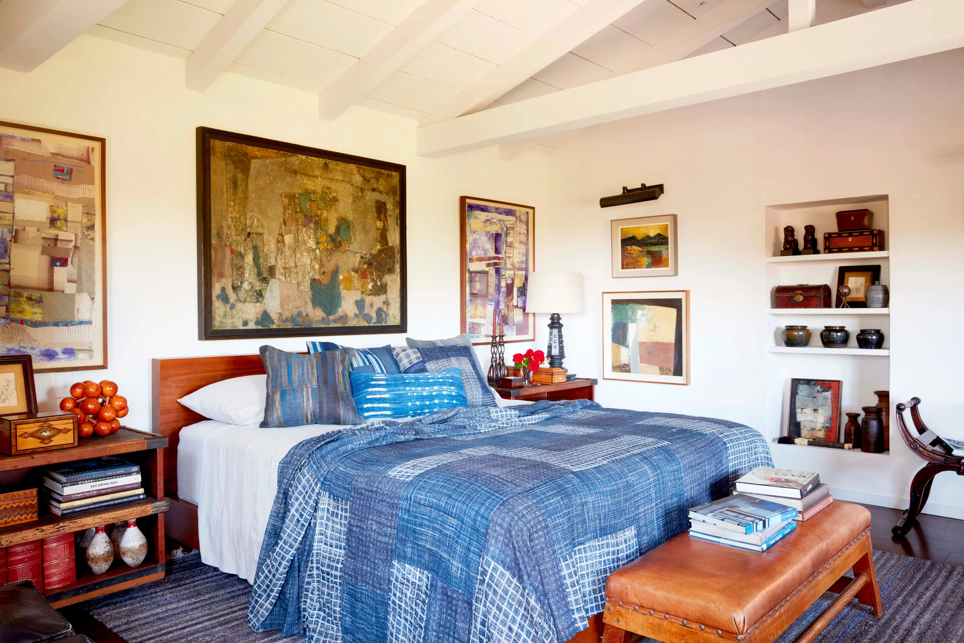 An eclectic bedroom featuring a bed styled with a blue patchwork quilt, a leather bench, and white exposed ceiling beams.