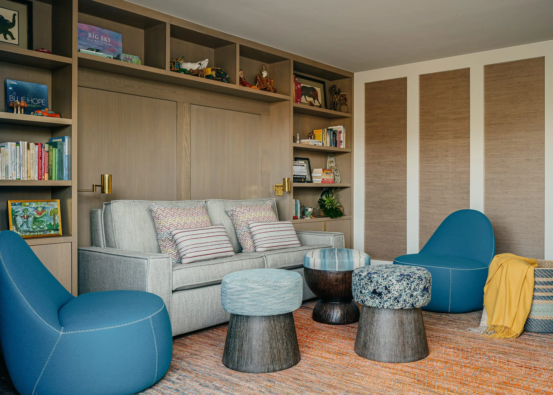 A family room seating area features a grey sofa set against a light wood built-in shelving unit, flanked by two blue armchairs on an orange patterned rug.