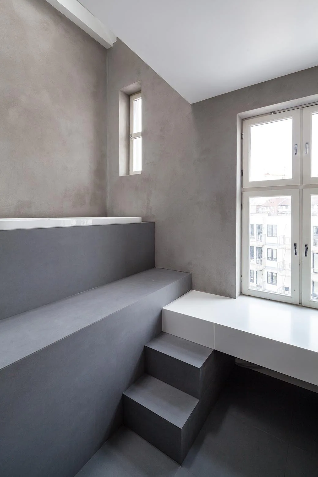 A minimalist grey bathroom featuring a white built-in bathtub on an elevated concrete platform with steps next to a window.